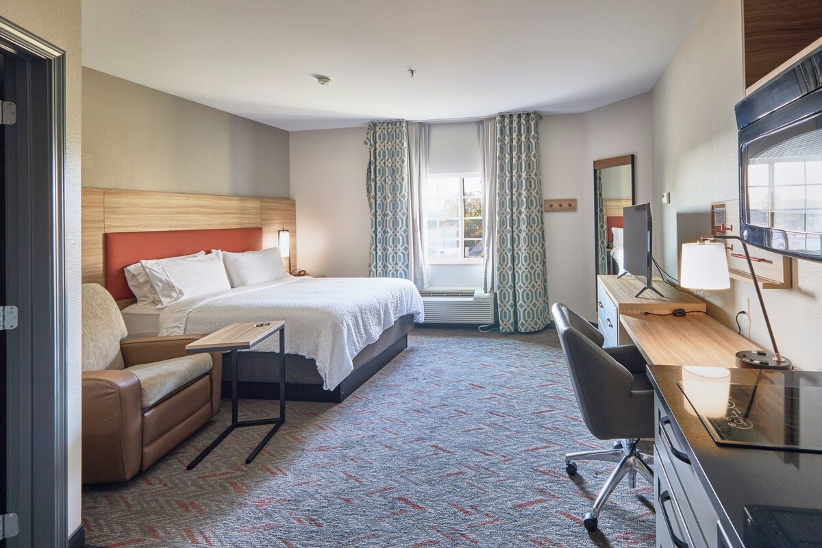 Ein Hotelzimmer mit Bett, Schreibtisch, Stuhl, einer Couch und einem großen Fenster mit Vorhängen.