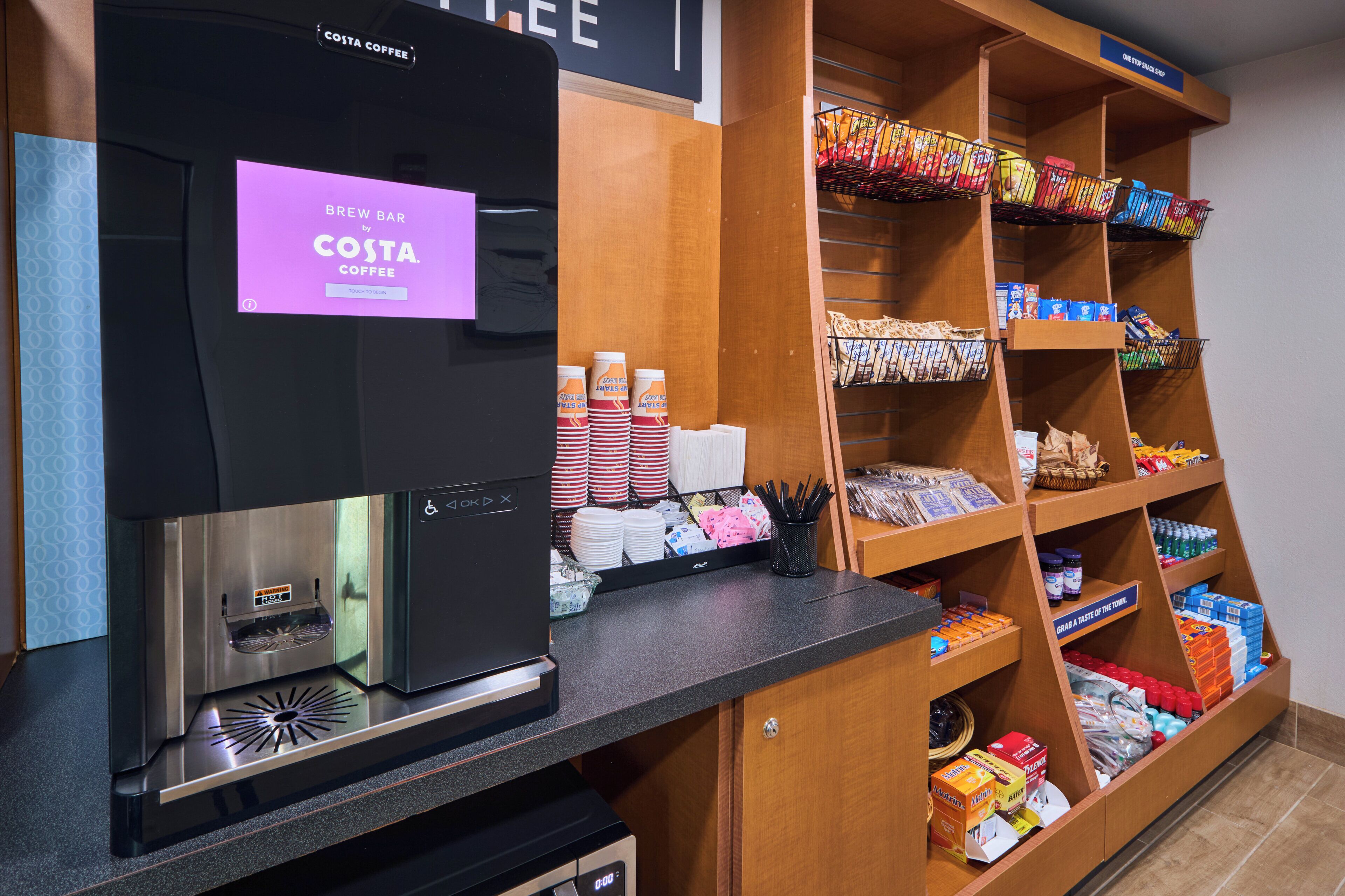 A coffee station with a branded coffee machine, a display of coffee cups, and shelves stocked with snacks and beverages.