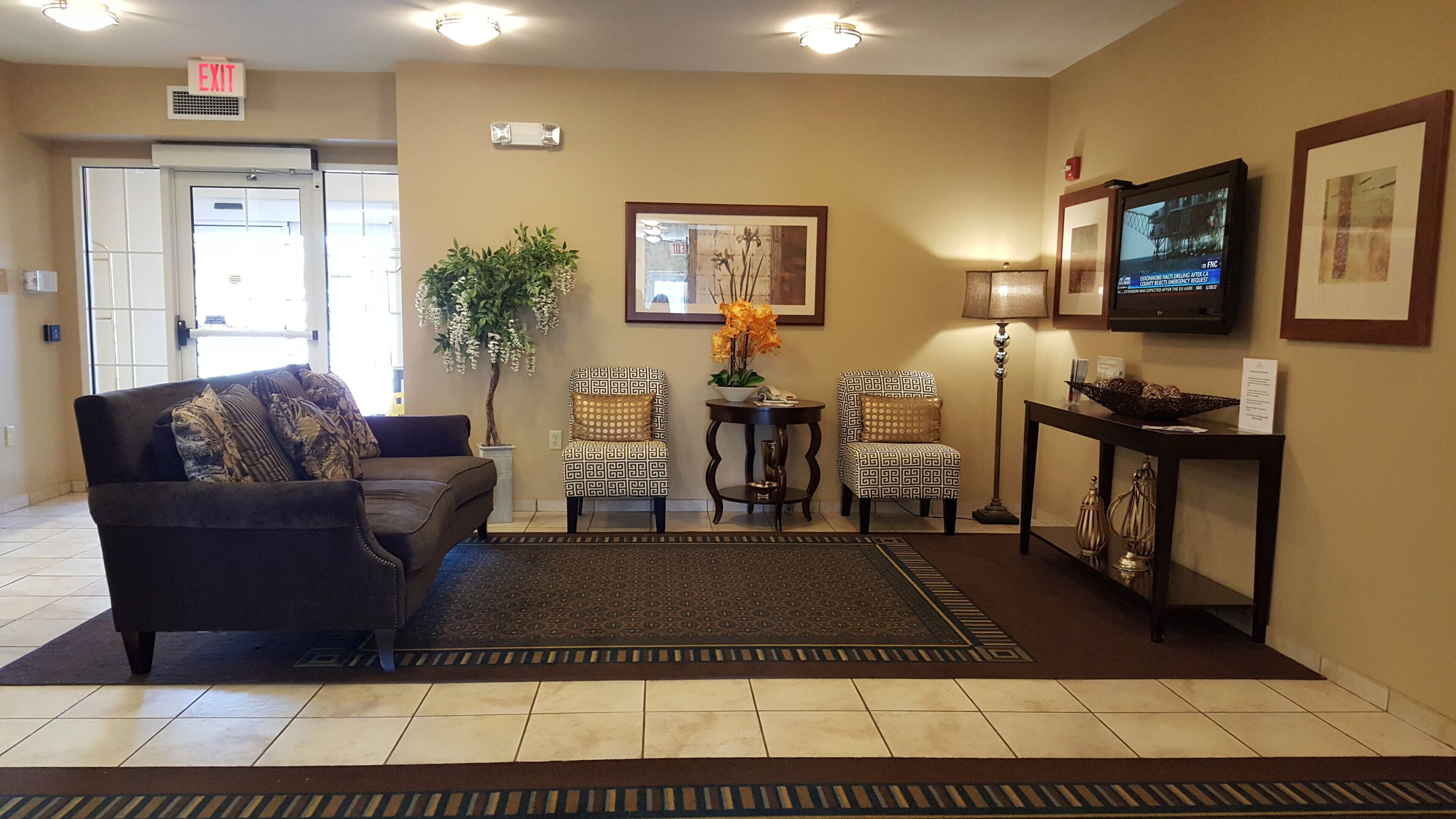 A hotel lobby with a sofa, two patterned chairs, a small table with a plant, a lamp, and a television on a stand.
