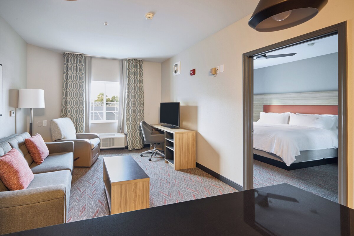A hotel room with a bed, a desk, a chair, a television, and a view of the sea through an open door.