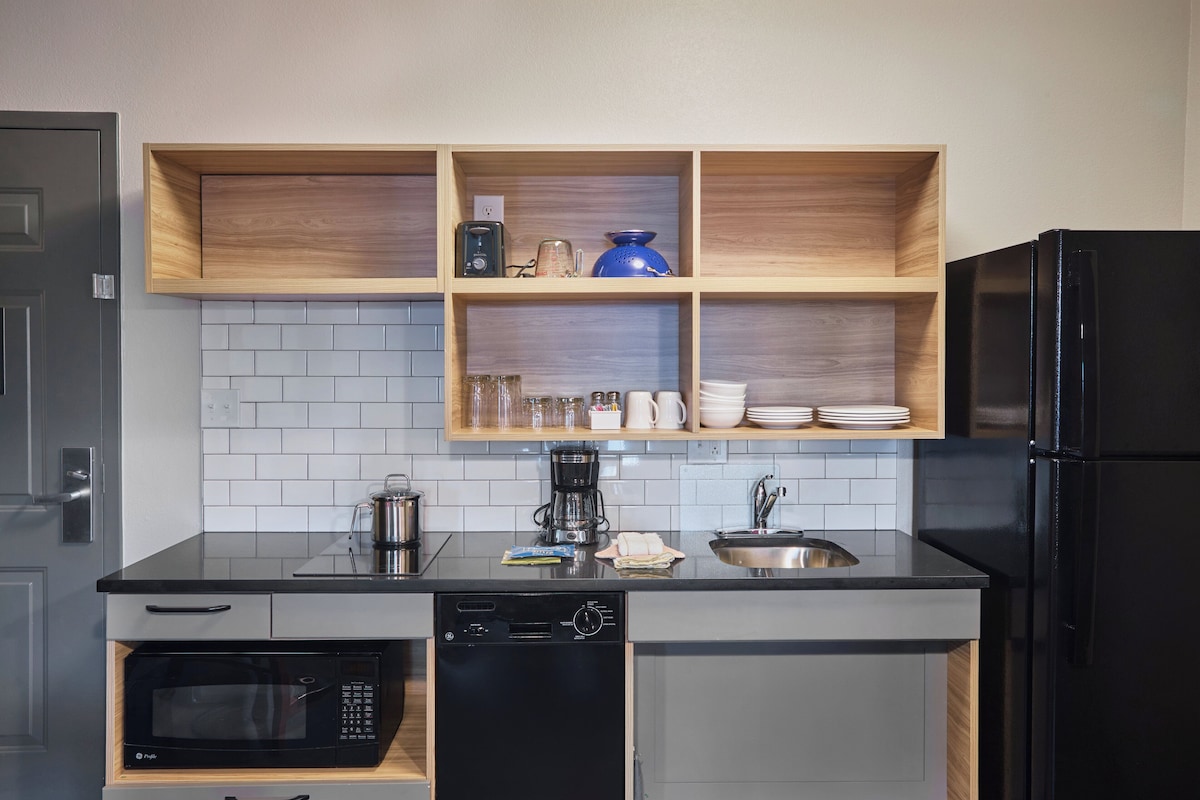 A compact kitchen with a black refrigerator, a microwave, a dishwasher, and wooden shelves above the countertop.