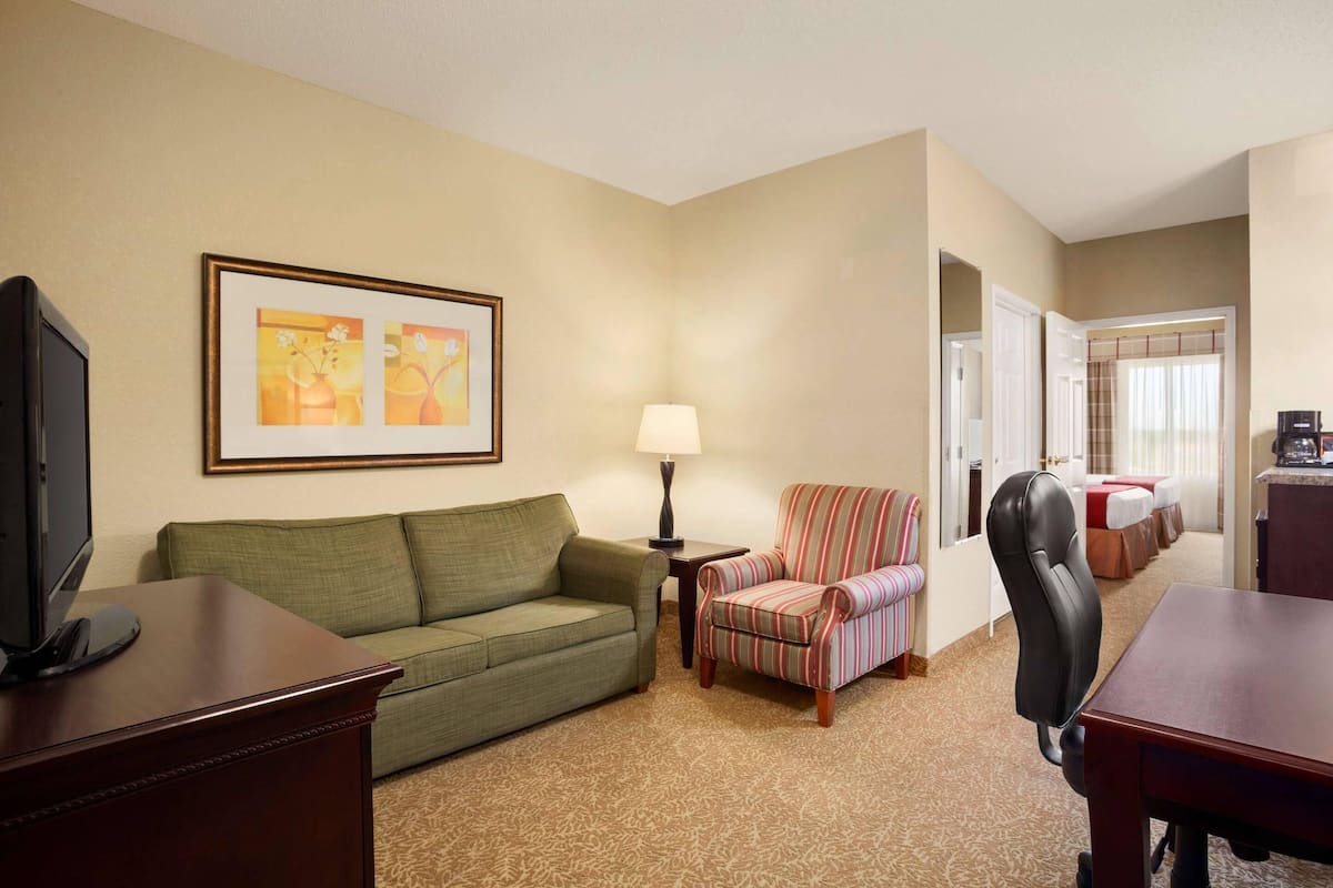 Habitación de hotel con un sofá verde, un sillón a rayas, un escritorio con una computadora, una lámpara y un cuadro en la pared.