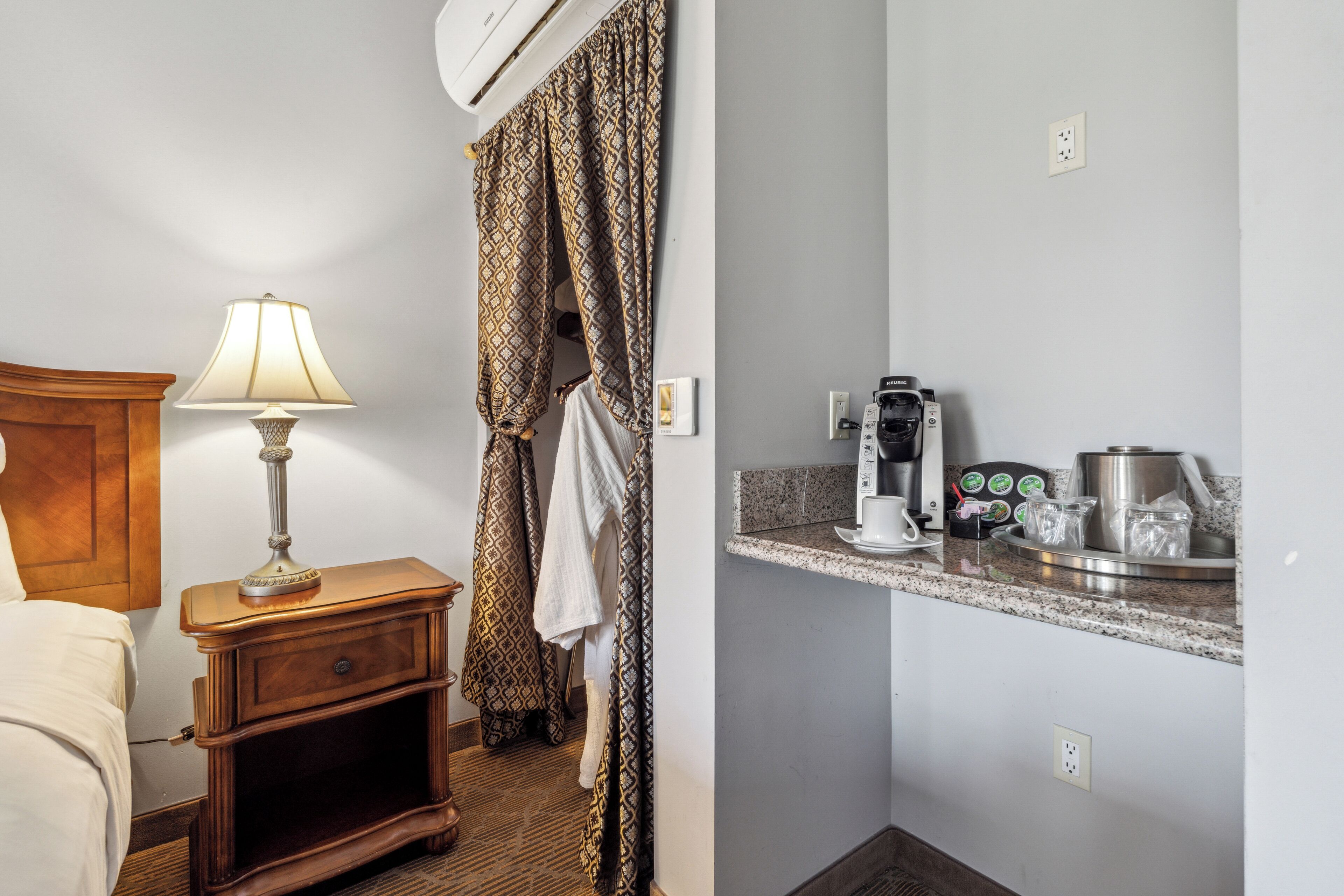 Une chambre d’hôtel avec une table de chevet en bois, une lampe, un rideau à motifs, une serviette suspendue, une kitchenette équipée d’une machine à café et un lit.