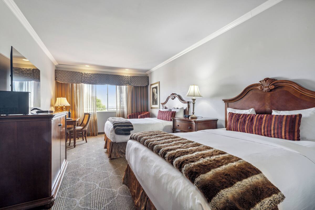 A hotel room with two beds, a desk, a chair, a TV, and a window with curtains.