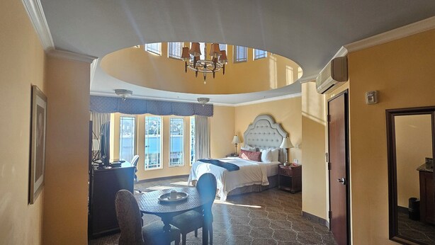 Una habitación de hotel con una cama grande, una mesa de comedor, una araña y vistas de la ciudad a través de las ventanas.