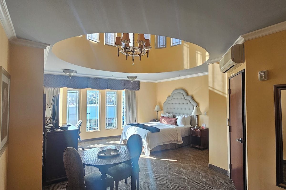 A hotel room with a large bed, a dining table, a chandelier, and a view of the city through the windows.