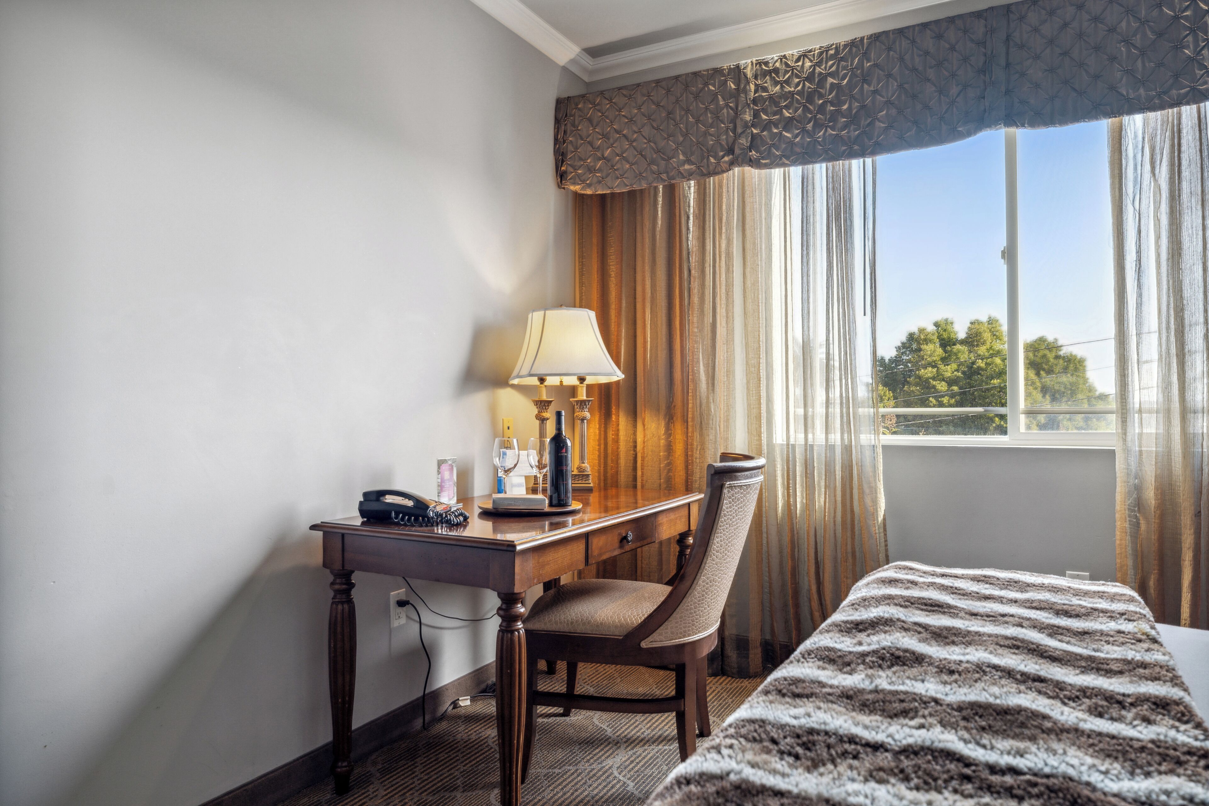 A hotel room with a bed, a desk with a lamp, a chair, and a view of trees through the window.