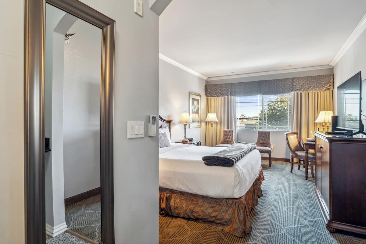 A hotel room with a large bed, two bedside tables with lamps, a desk with a chair, a television, and a window with curtains.