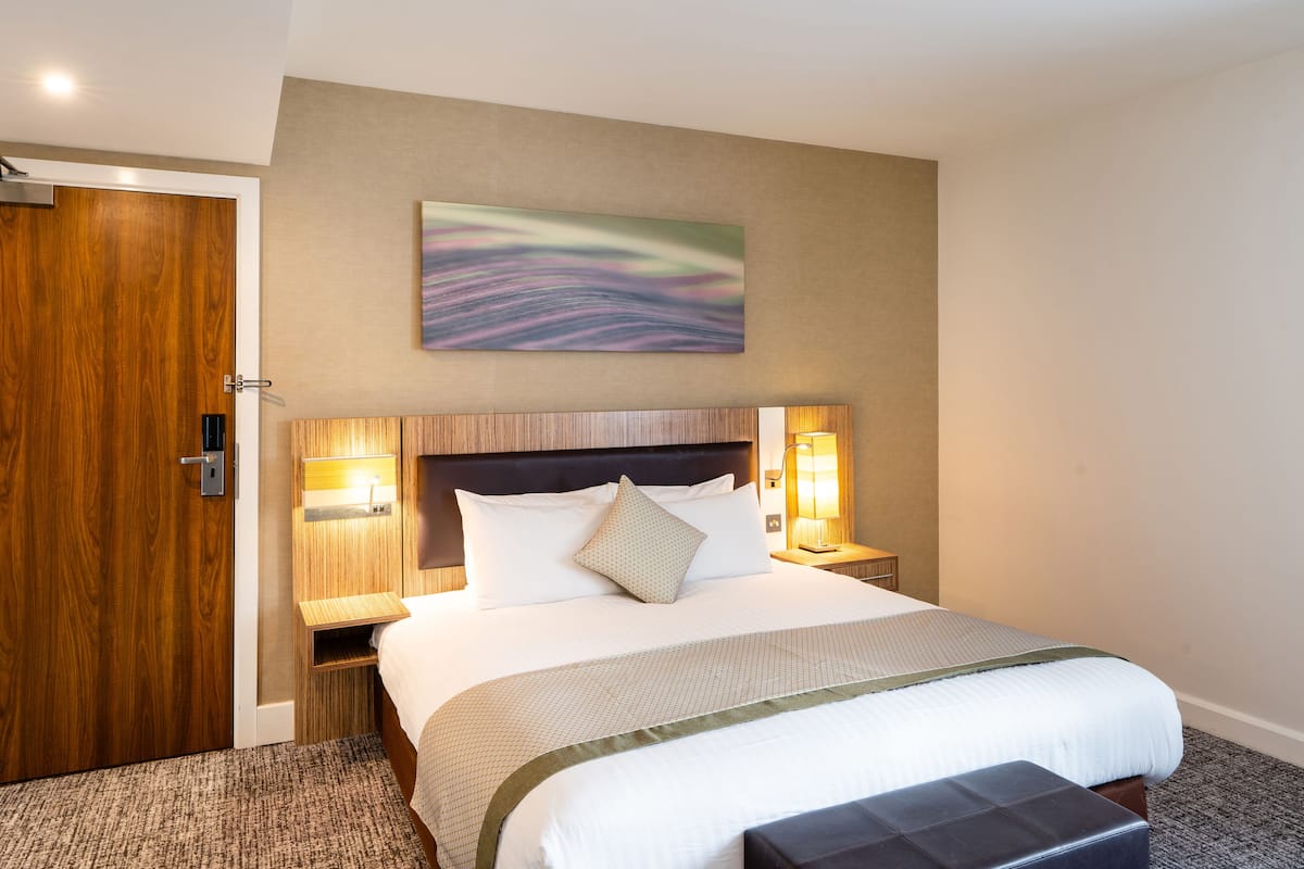 A hotel room with a large bed, a wooden door, a headboard with wall-mounted lights, and a painting on the wall.