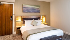 A hotel room with a large bed, a wooden door, a headboard with wall-mounted lights, and a painting on the wall.
