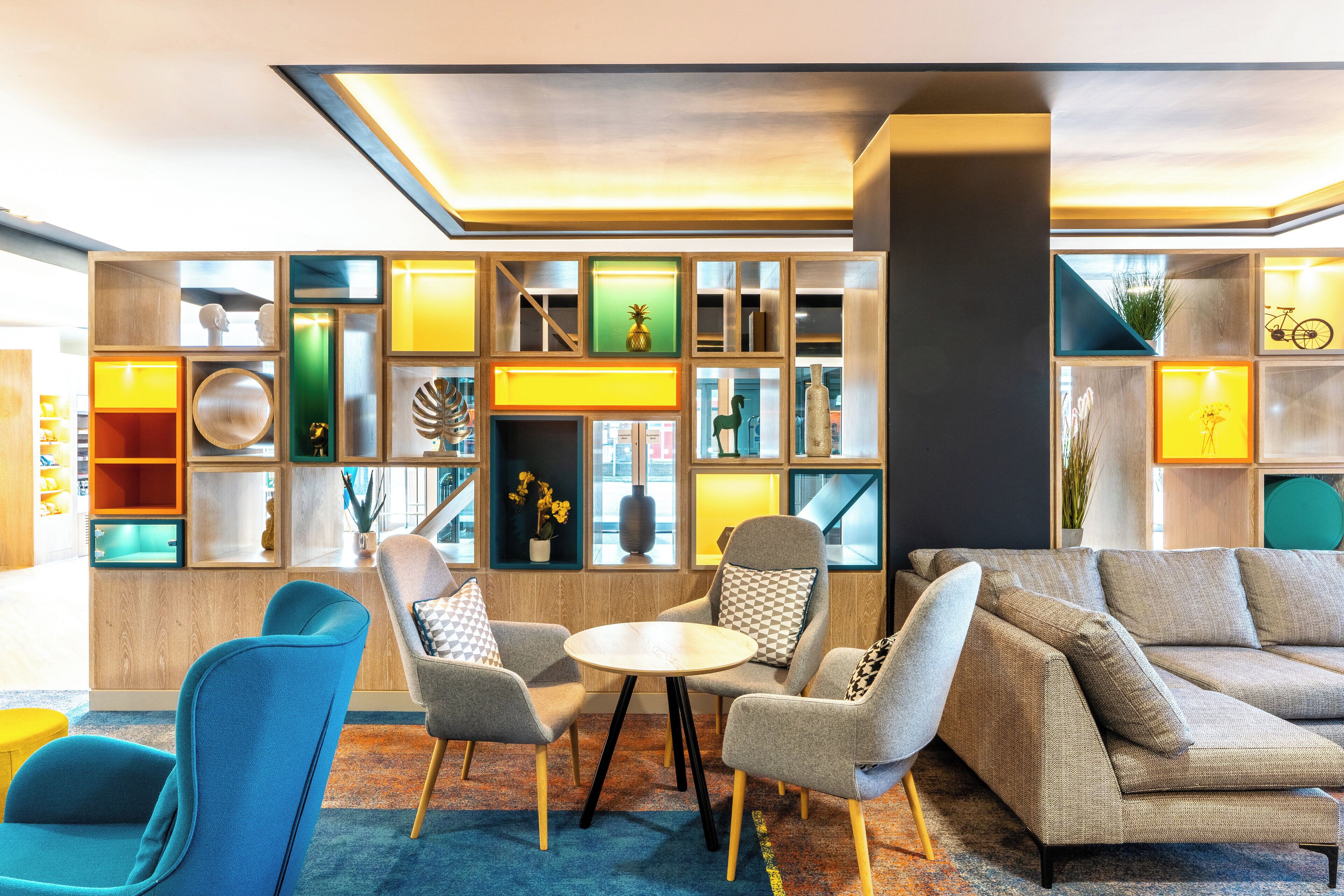 A modern lounge area with a sectional sofa, armchairs, and a small round table. There are shelves with various decorative items and a large window providing natural light.