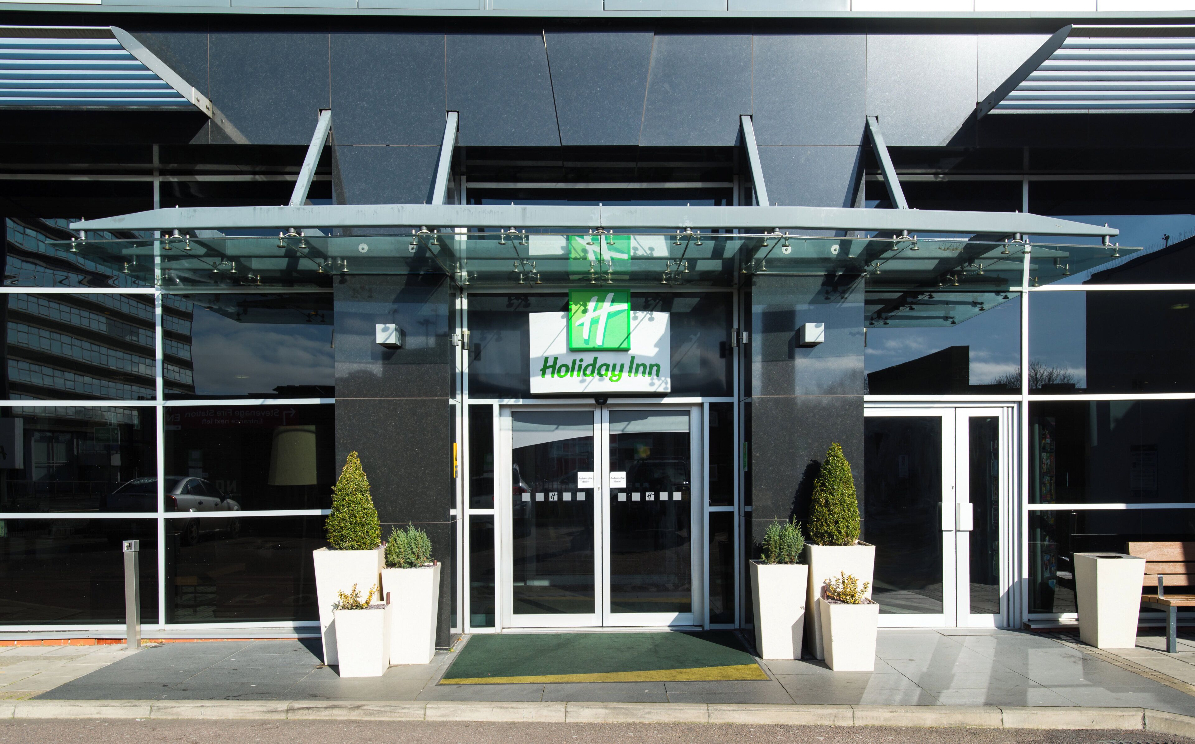 Der Eingang eines Holiday Inn Hotels mit einer Glasüberdachung, einem grünen Schild und Pflanzkübeln.