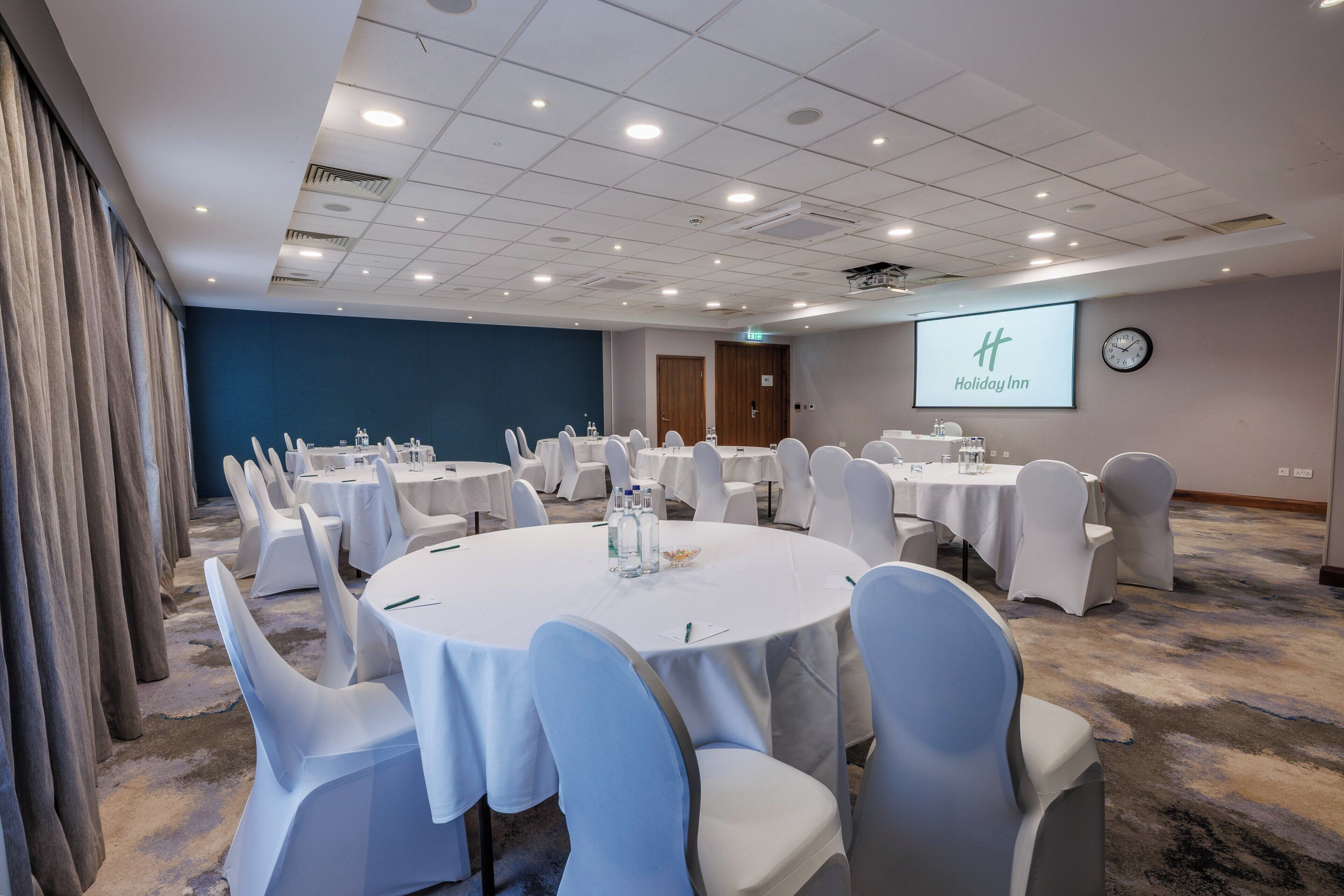 A conference room with round tables, white chairs, a clock, and a screen displaying the Holiday Inn logo.
