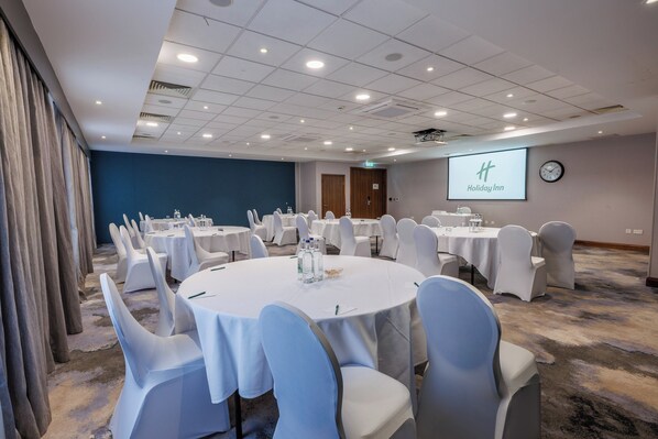 A conference room with round tables, white chairs, a clock, and a screen displaying the Holiday Inn logo.