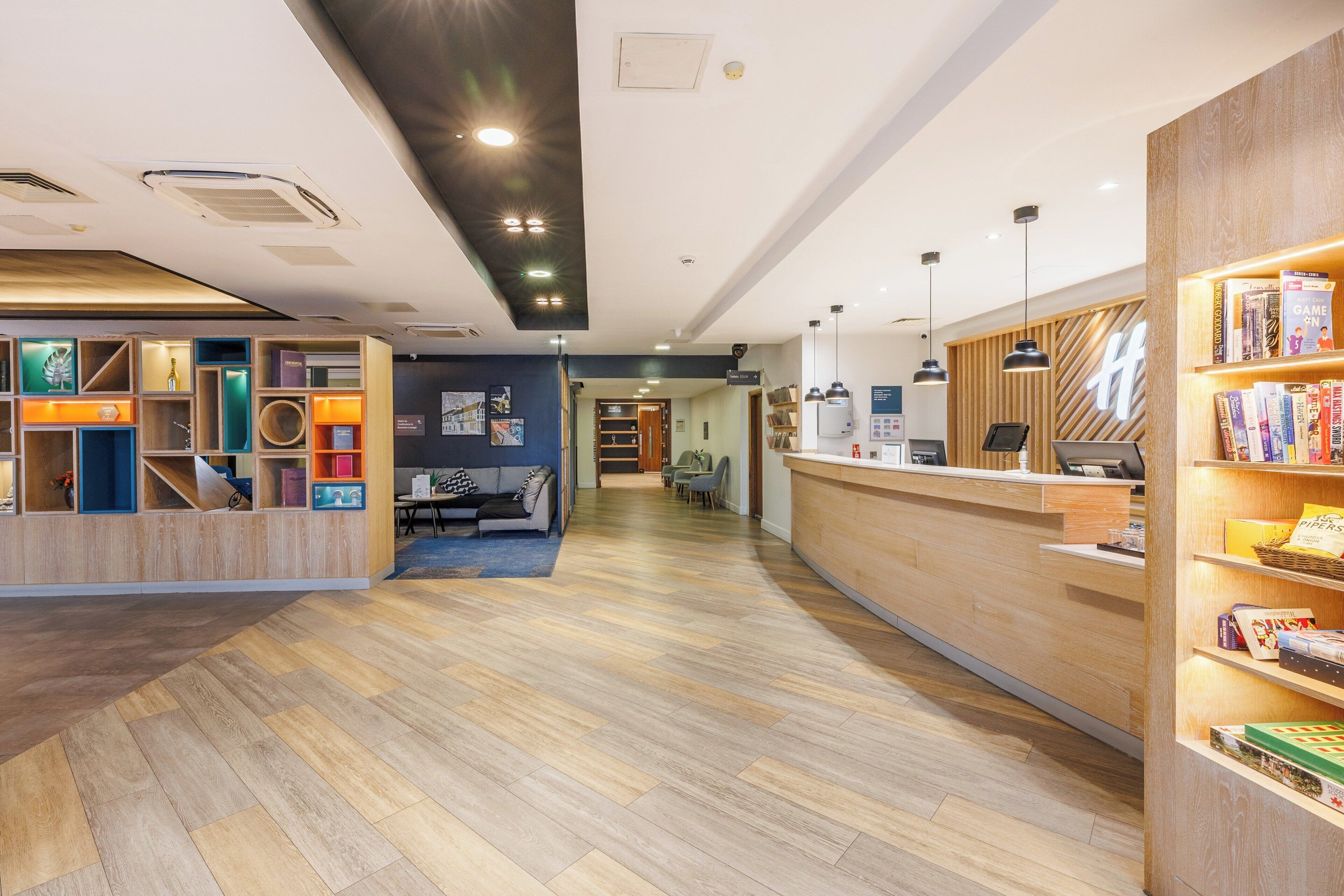 A modern reception area with a wooden reception desk, a seating area with a sofa and chairs, and a bookshelf with various books.