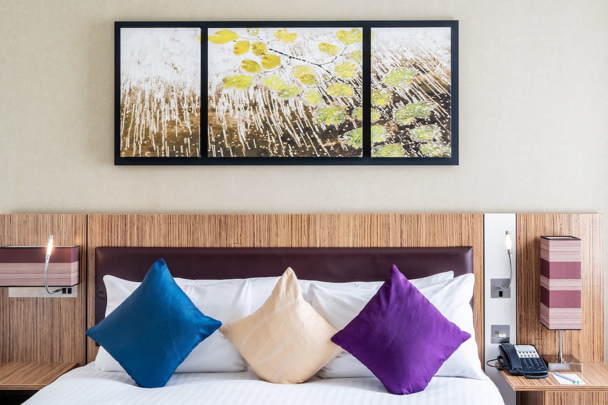 A hotel room with a bed, pillows, a bedside table with a phone, and a wall-mounted artwork.