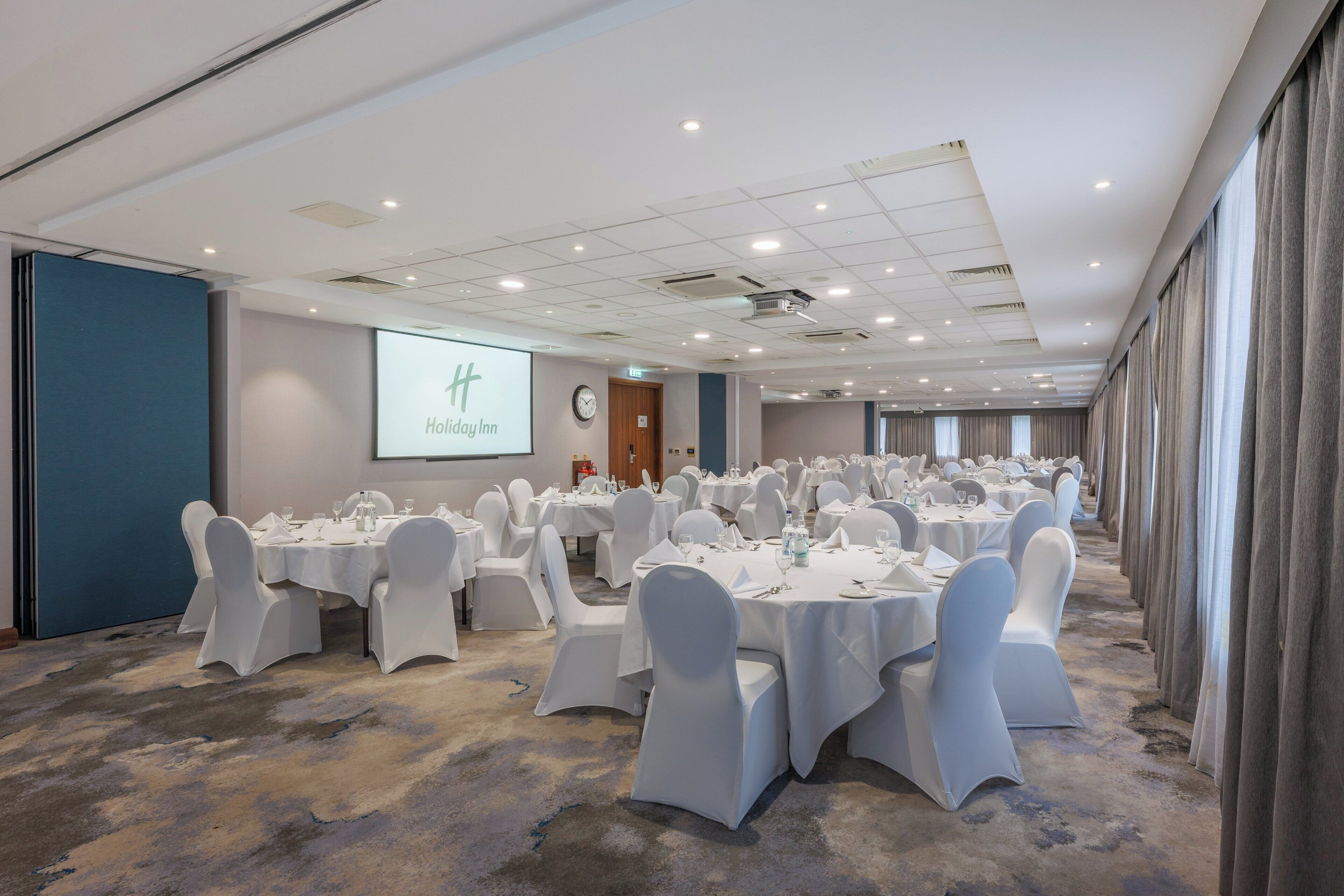 A conference room with rows of tables and chairs, a projector screen displaying 'Holiday Inn', and a clock on the wall.