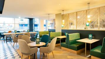 A modern dining area with green booths and round tables, featuring a geometric patterned floor and a view of the city through large windows.