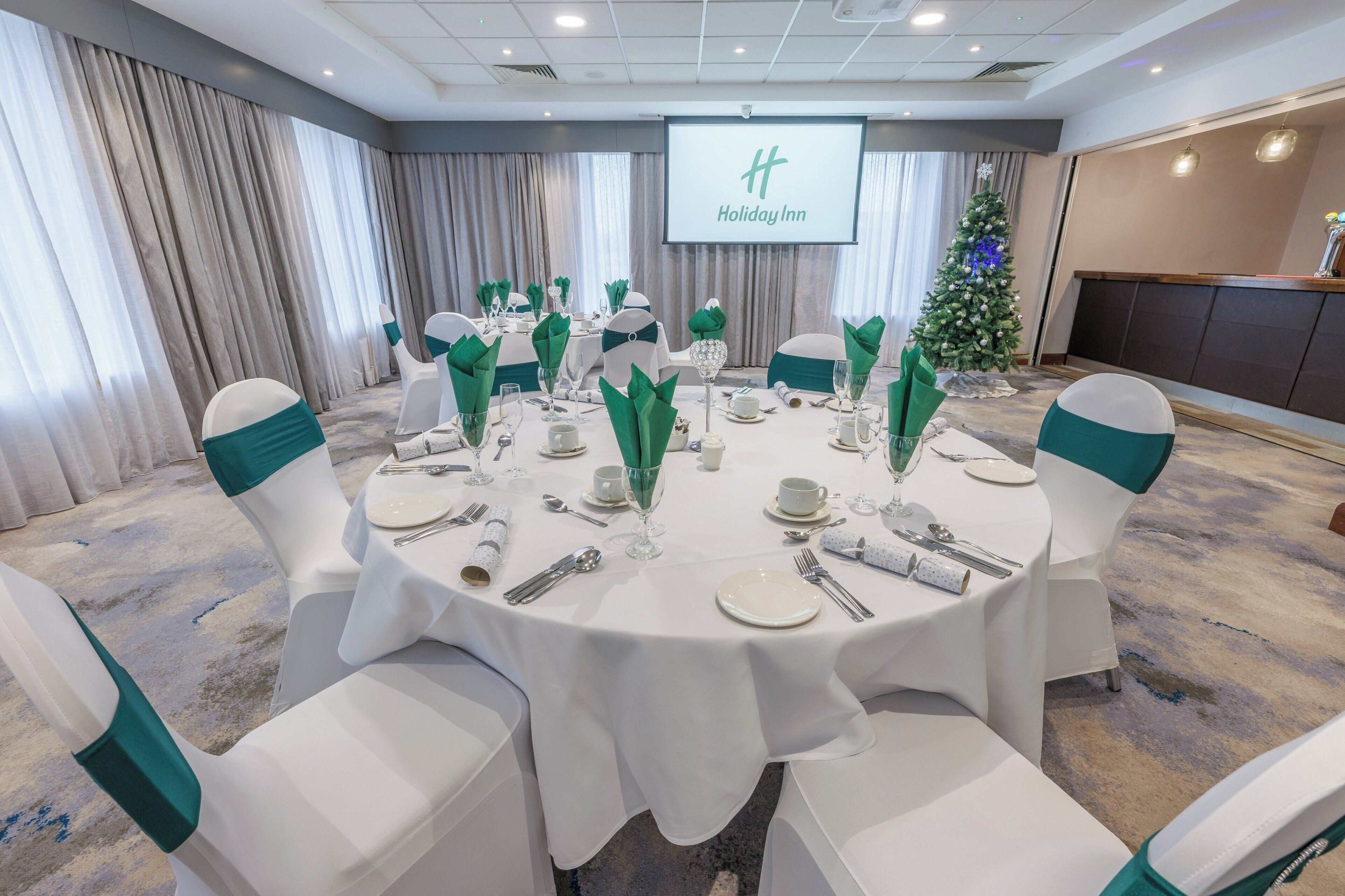 A conference room with a round table set for a meeting, white chairs with green sashes, a Christmas tree, and a 'Holiday Inn' sign on the wall.
