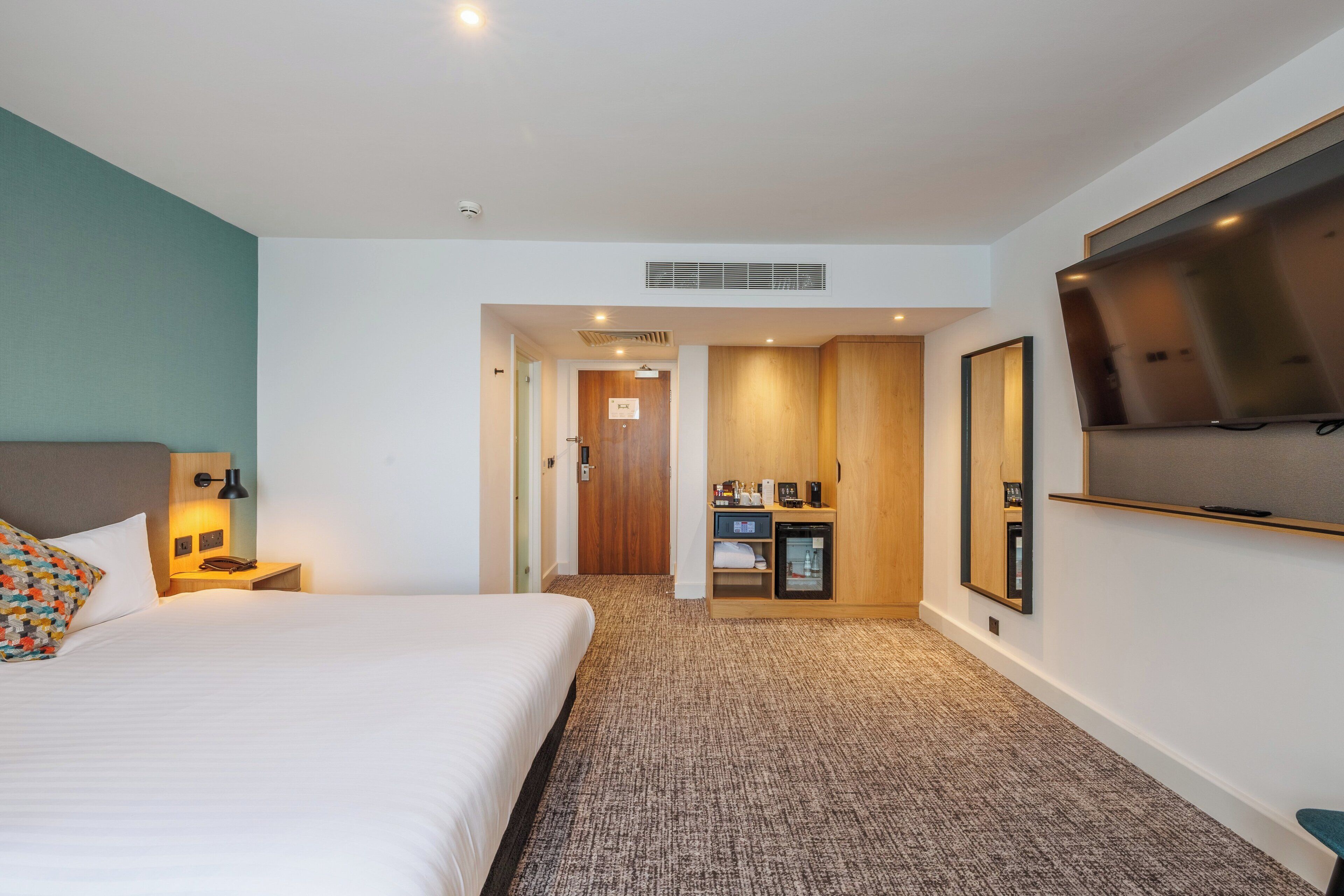 A hotel room with a bed, a desk with a lamp, a television, and a minibar.