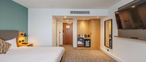 A hotel room with a bed, a desk with a lamp, a television, and a minibar.