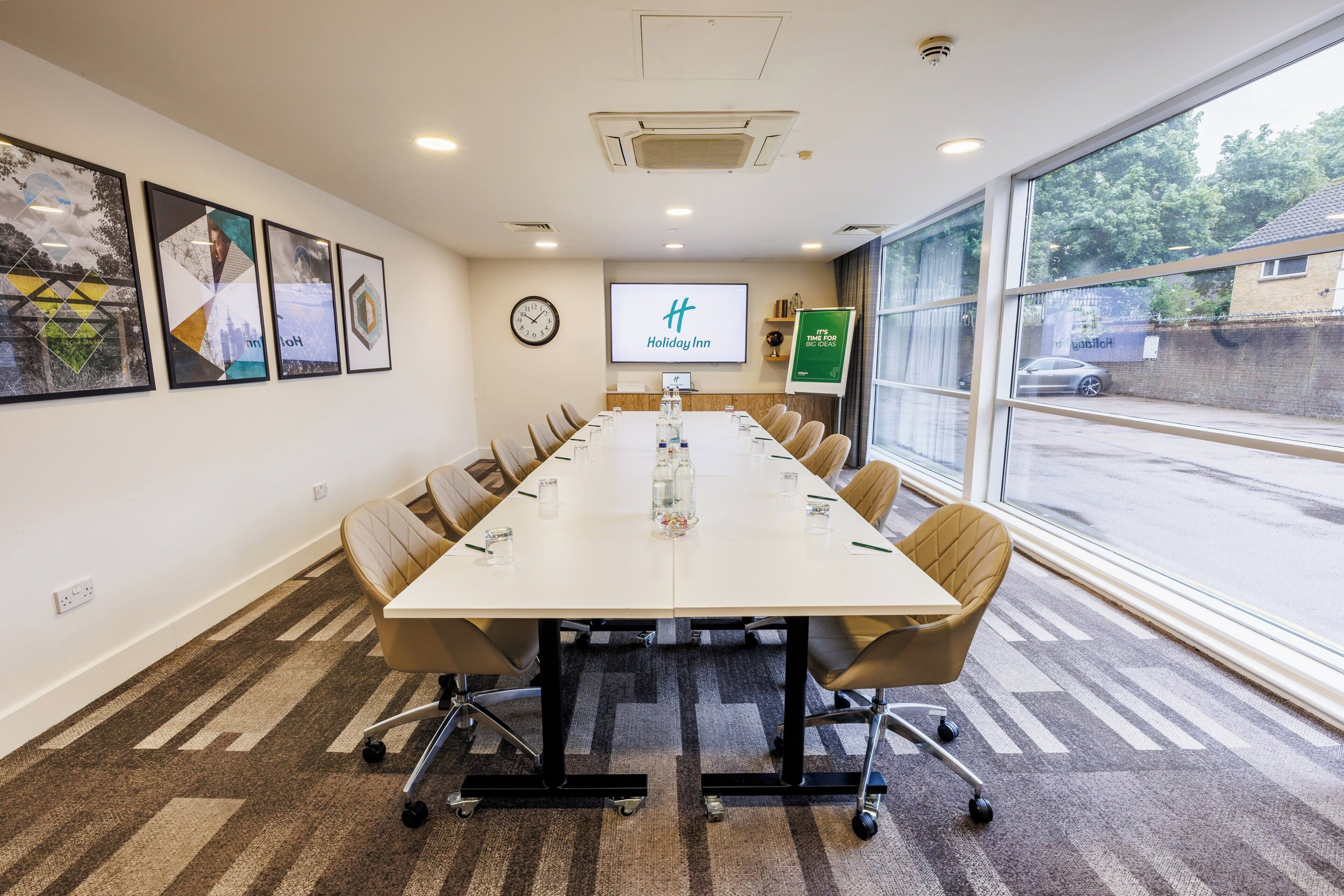 A conference room with a long table, chairs, a clock, a screen displaying 'Holiday Inn', and a window with a view of the outside.