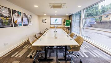 A conference room with a long table, chairs, a clock, a screen displaying 'Holiday Inn', and a window with a view of the outside.