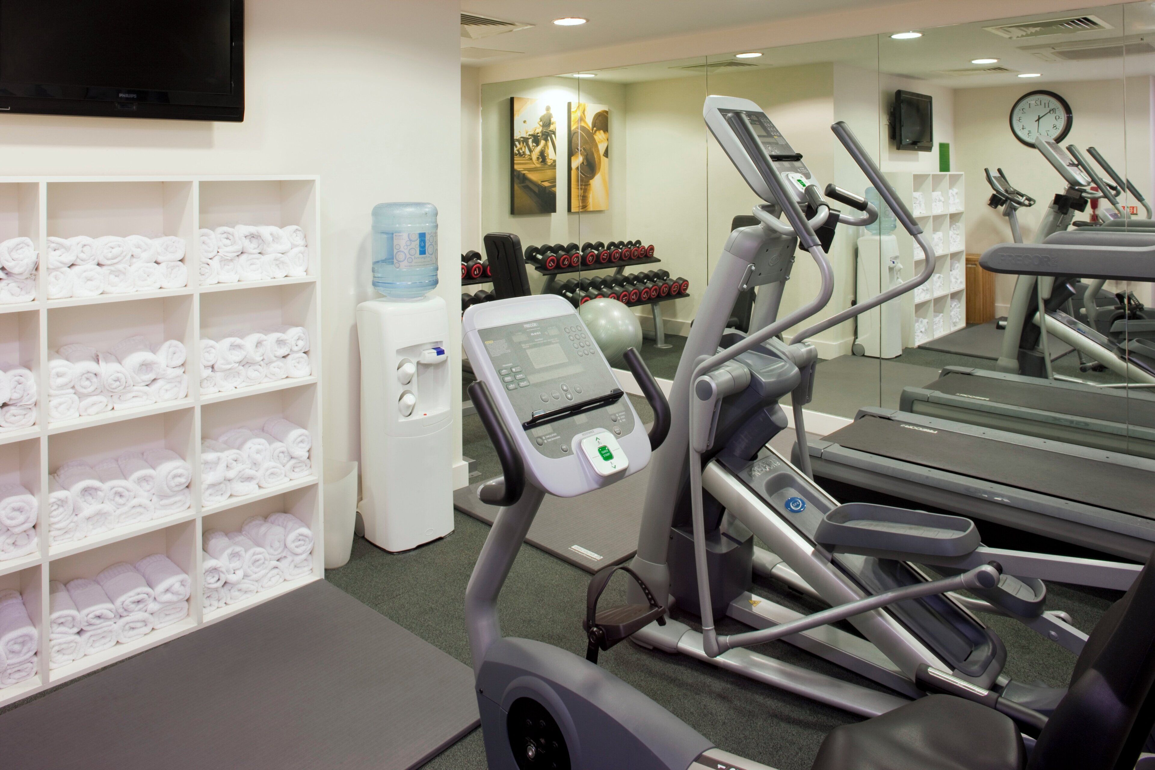 Un gimnasio con equipo de ejercicio, dispensadores de agua y toallas dobladas en estantes.