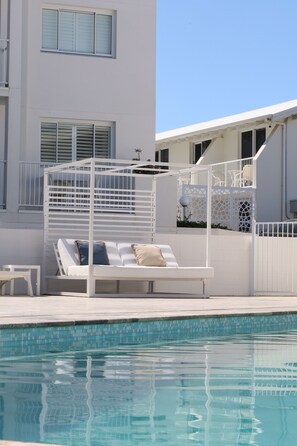 Zona junto a la piscina con mobiliario exterior blanco, que incluye una pérgola y sillones reclinables.