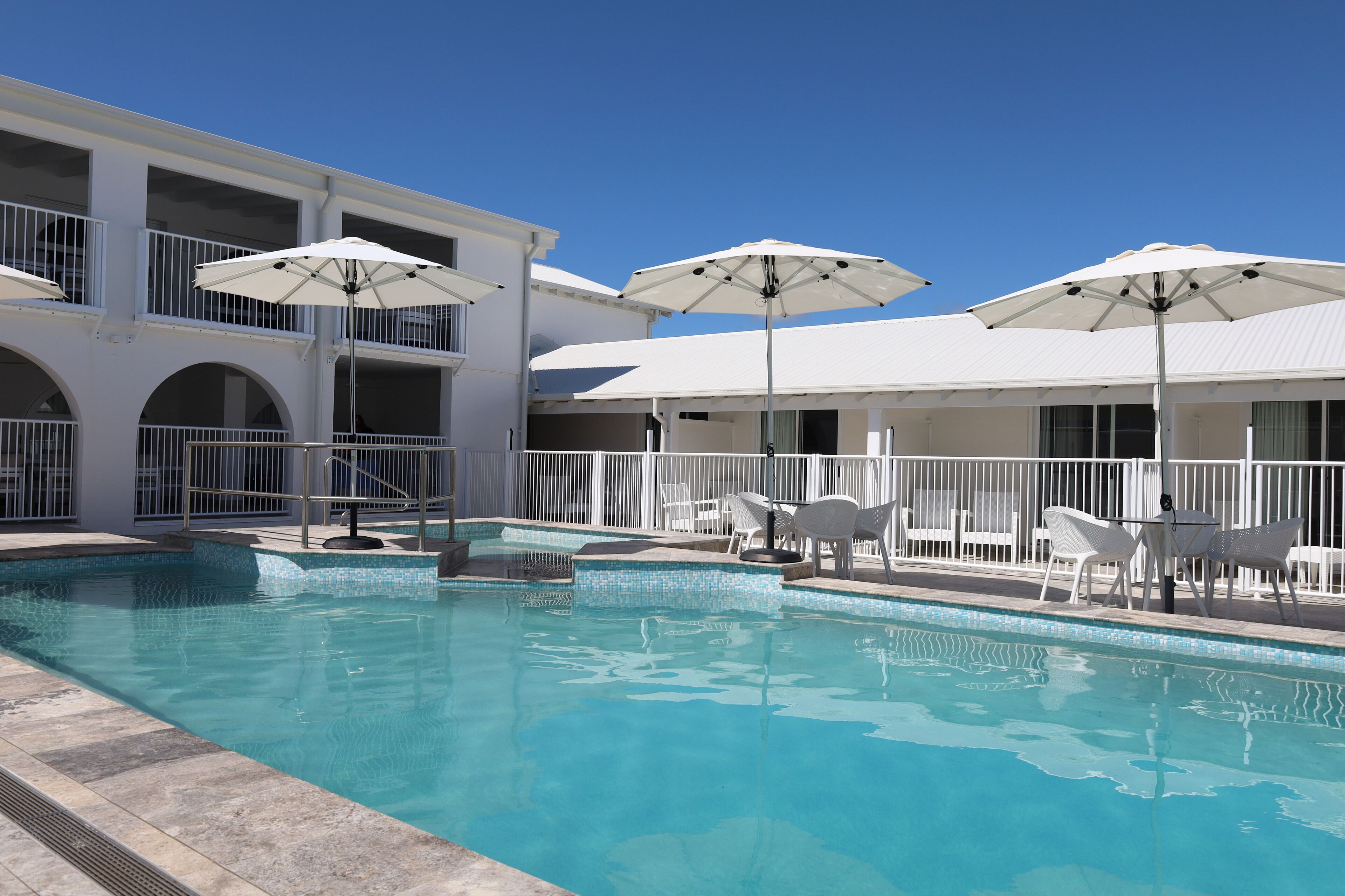 Una piscina con sombrillas y sillas blancas, rodeada por un edificio blanco con balcones.