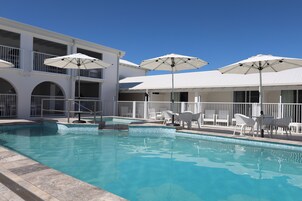 Una piscina con sombrillas y sillas blancas, rodeada por un edificio blanco con balcones.