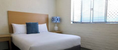 Una habitación de hotel con una cama grande, una mesita de noche con lámpara y una ventana con persianas.