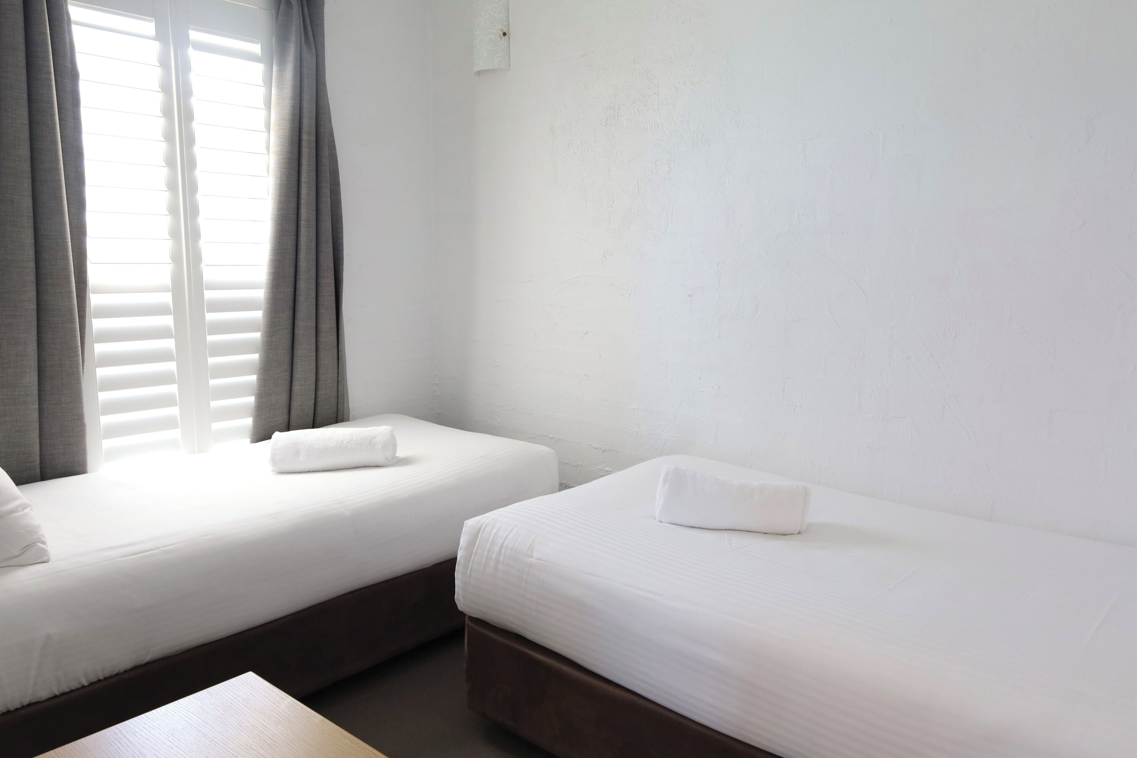 Two single beds with white linens and pillows, a wooden nightstand, and a window with grey curtains.