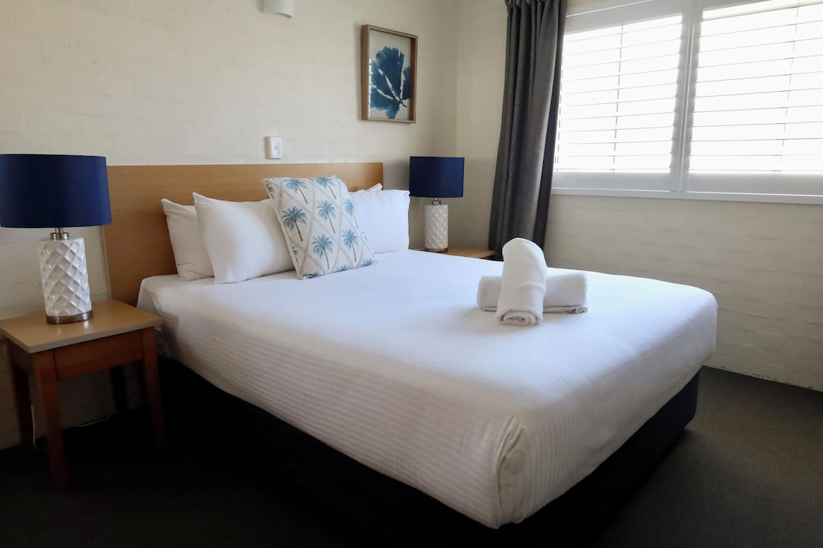 Een net opgemaakt bed met witte lakens en een blauw kussen, twee bedlampen, een kunstwerk in een lijst aan de muur, en een raam met gordijnen.