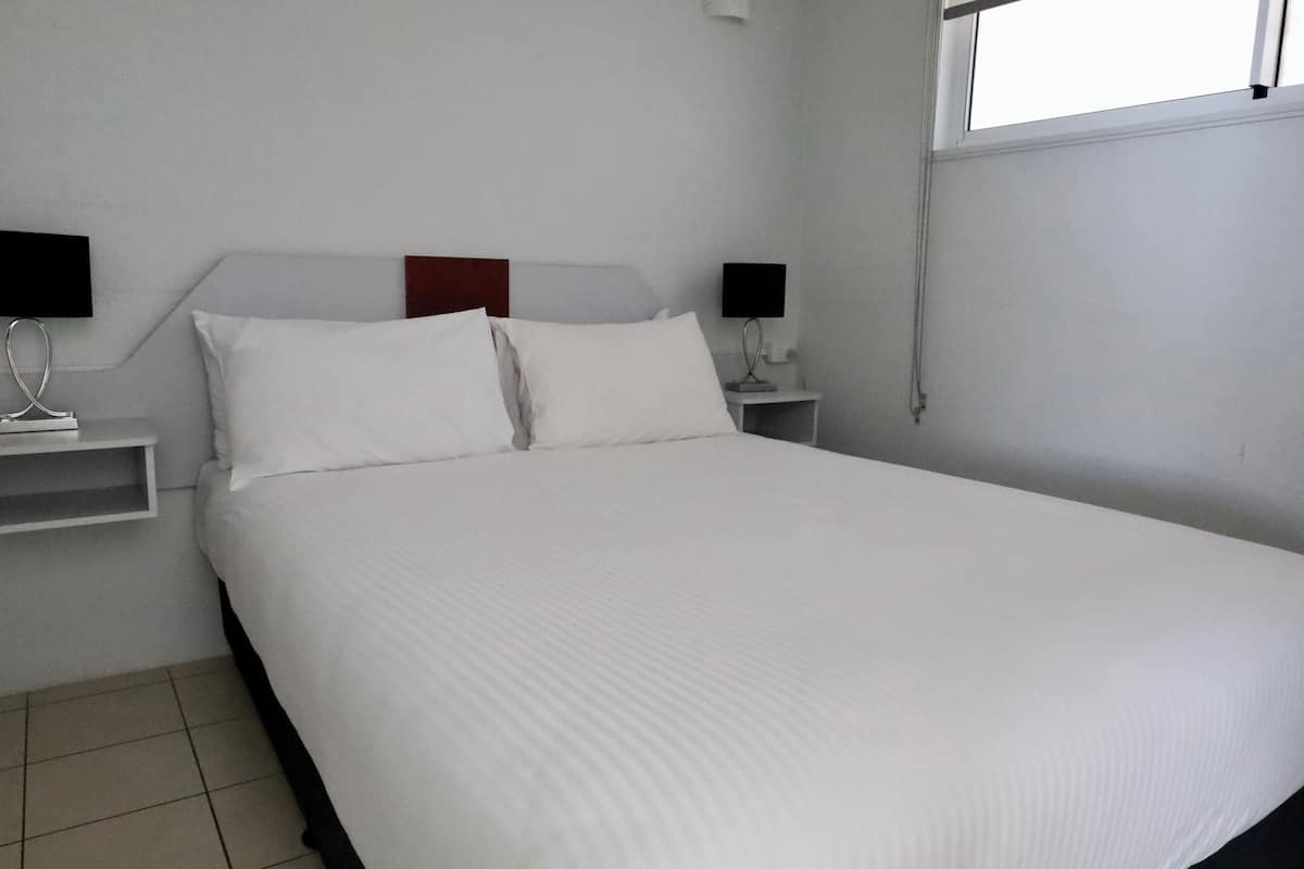 Een net opgemaakt bed met witte lakens in een eenvoudige kamer met witte wanden en een raam.
