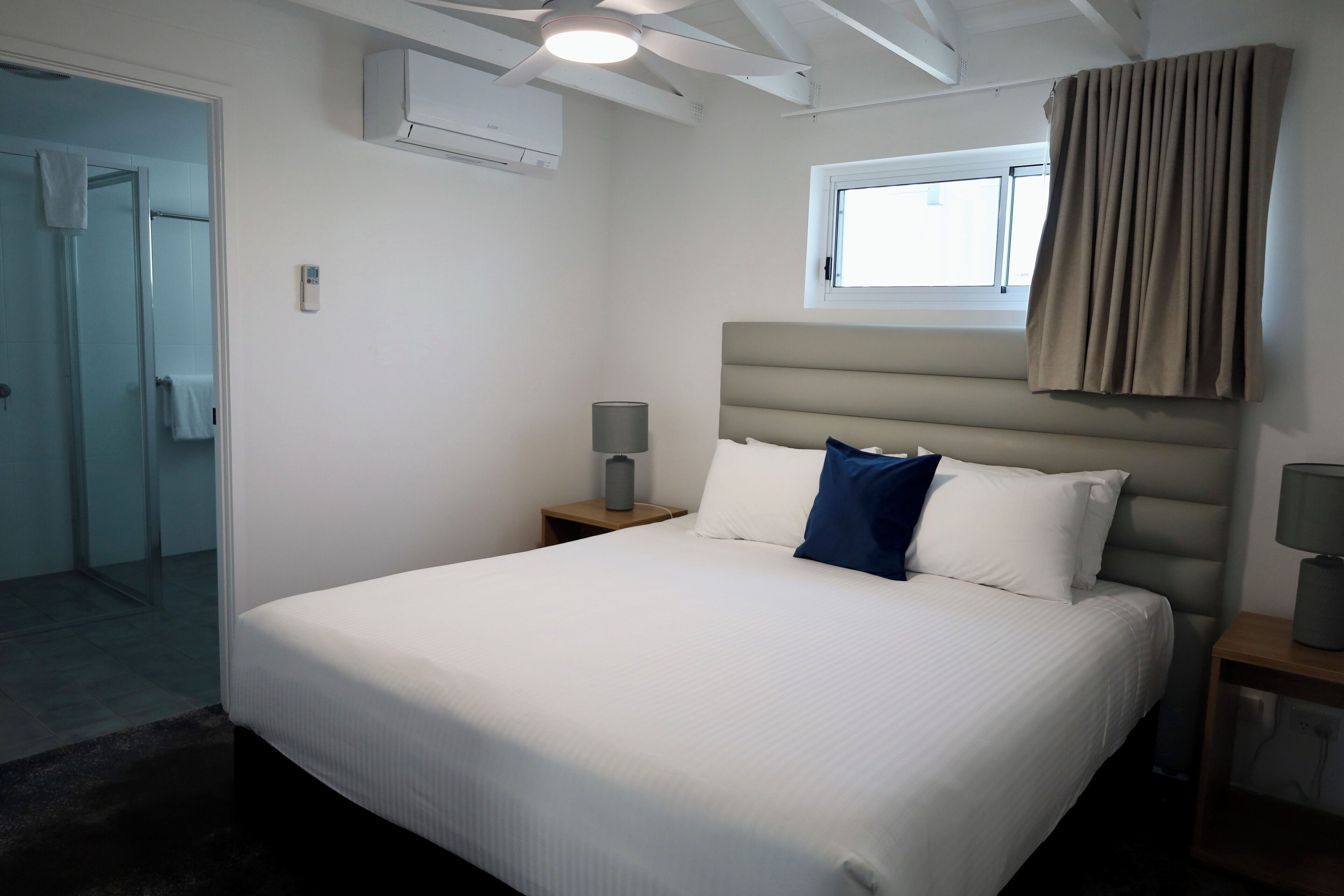 Une chambre d’hôtel avec un lit, une fenêtre avec des rideaux, un ventilateur de plafond et un climatiseur fix é au mur.