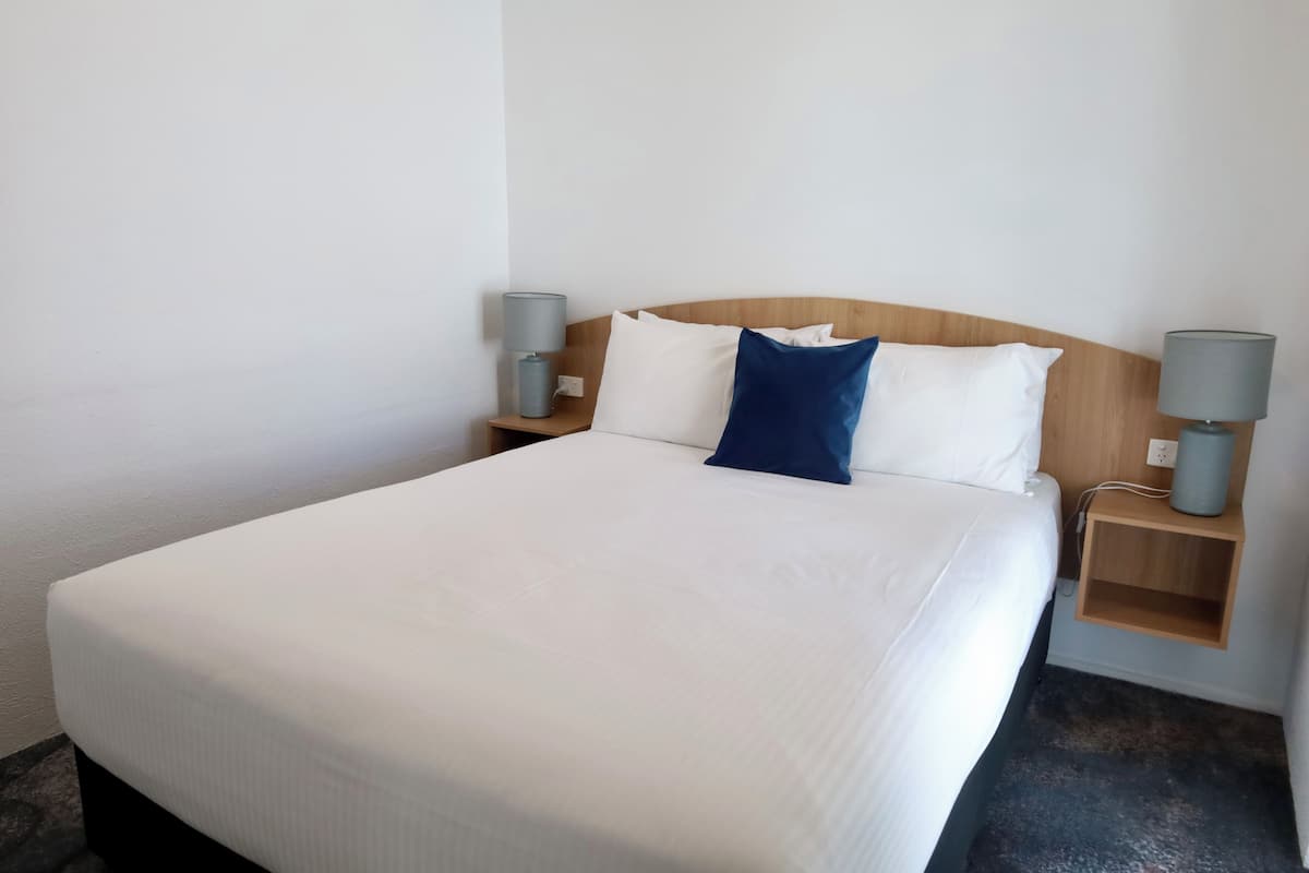 Een netjes opgemaakt bed met witte lakens en een blauw kussen, omringd door twee nachtkastjes met lampen, in een eenvoudige kamer met witte muren.