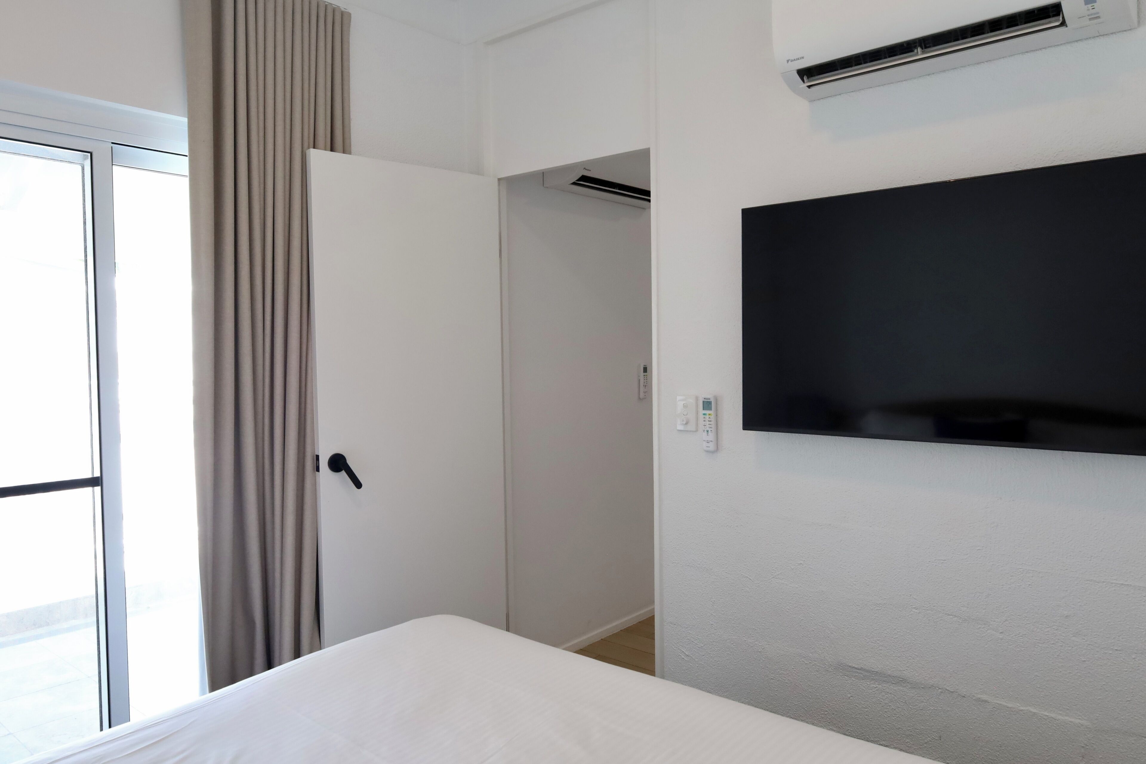 Una habitación de hotel con cama, televisión montada en la pared, aire acondicionado, una puerta y una ventana con cortinas.
