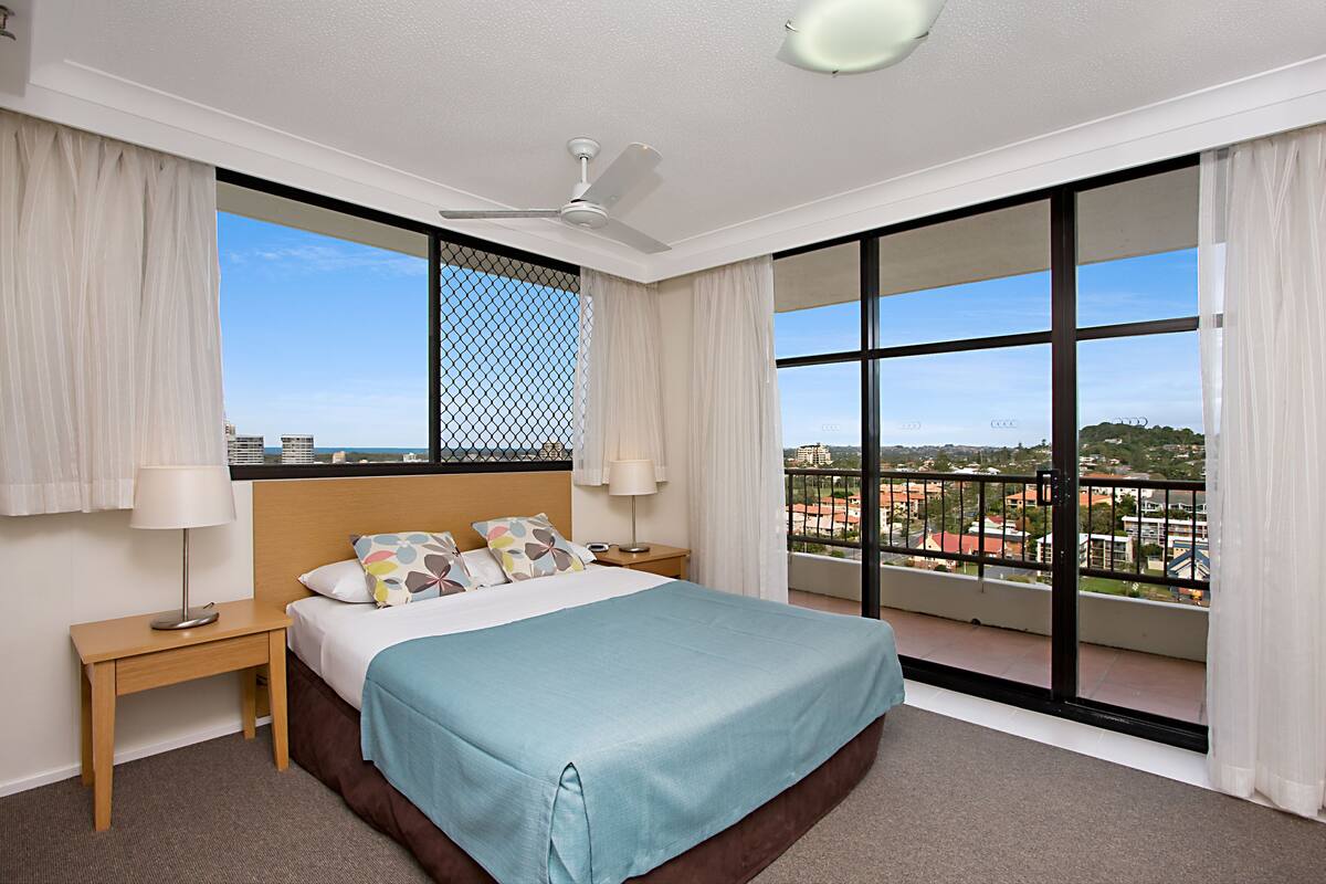 A hotel room with a large bed, two bedside tables, a ceiling fan, and a balcony with a view of the city.