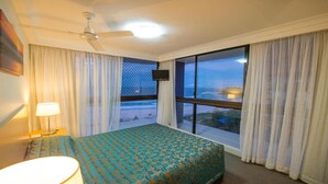 Une chambre d’hôtel équipée d’un lit, d’une télévision, d’un ventilateur de plafond et d’une grande fenêtre donnant sur la plage.