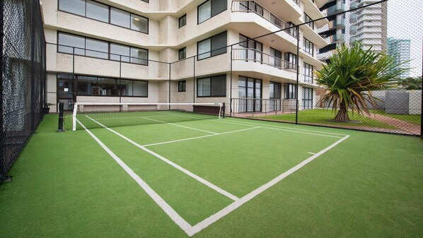 A tennis court with a net, surrounded by a chain-link fence, and adjacent to a multi-story building with balconies.