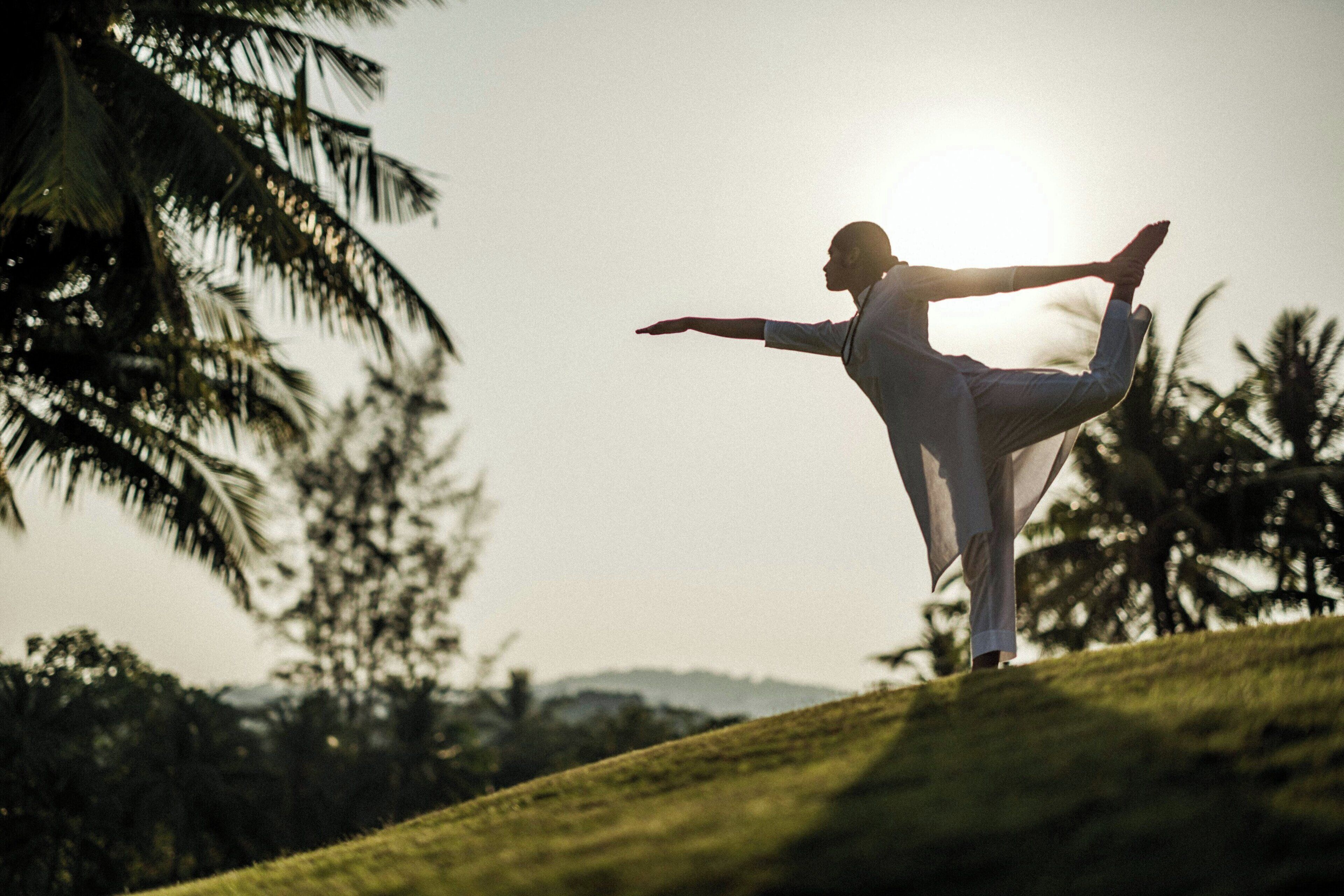 Une personne effectue une posture de yoga sur une pente herbeuse, avec des palmiers et un ciel dégagé en arrière-plan.