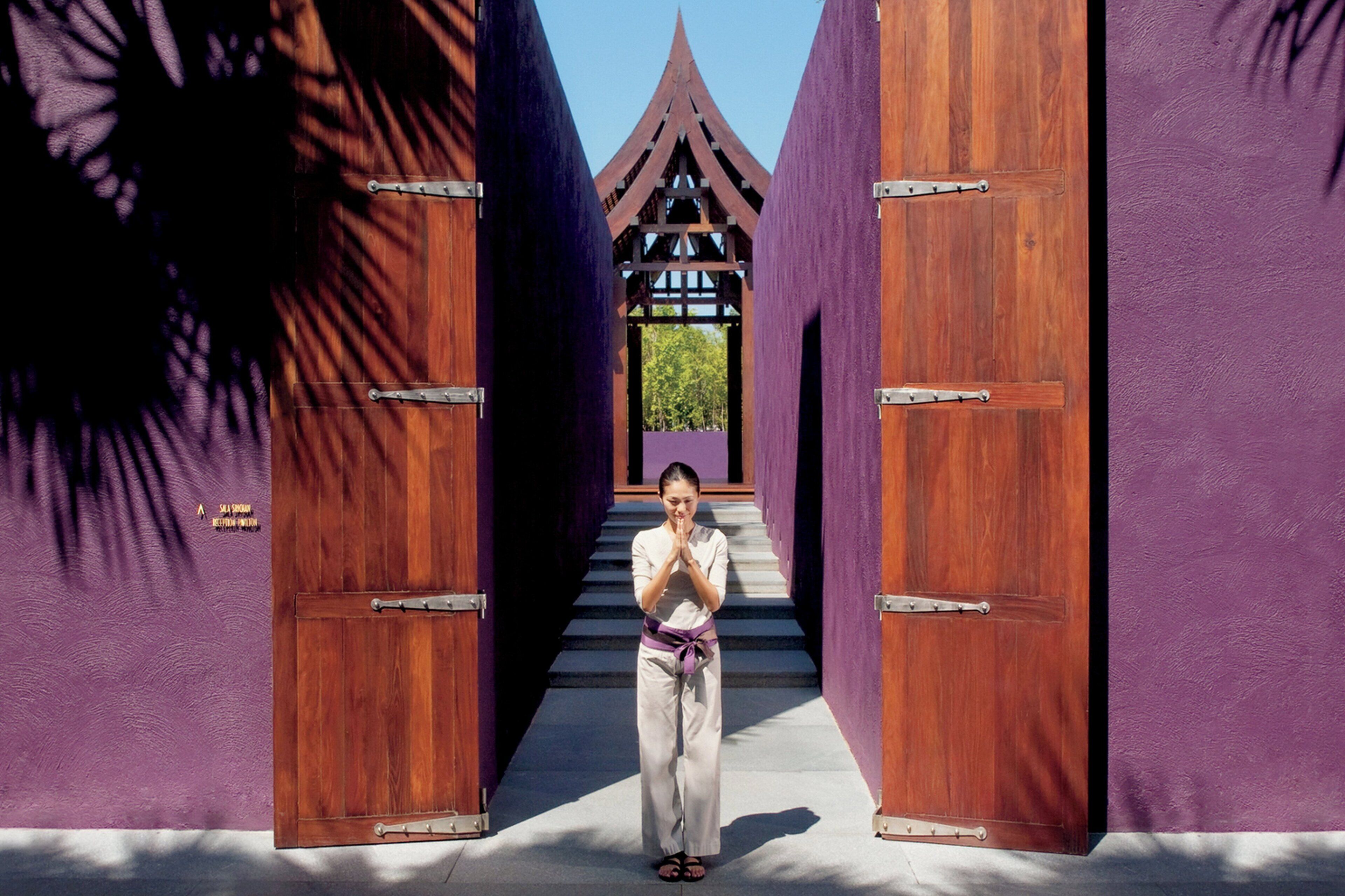 Eine Person steht zwischen zwei großen Holztüren, im Hintergrund eine violette Wand und ein traditionelles Dachgebilde.