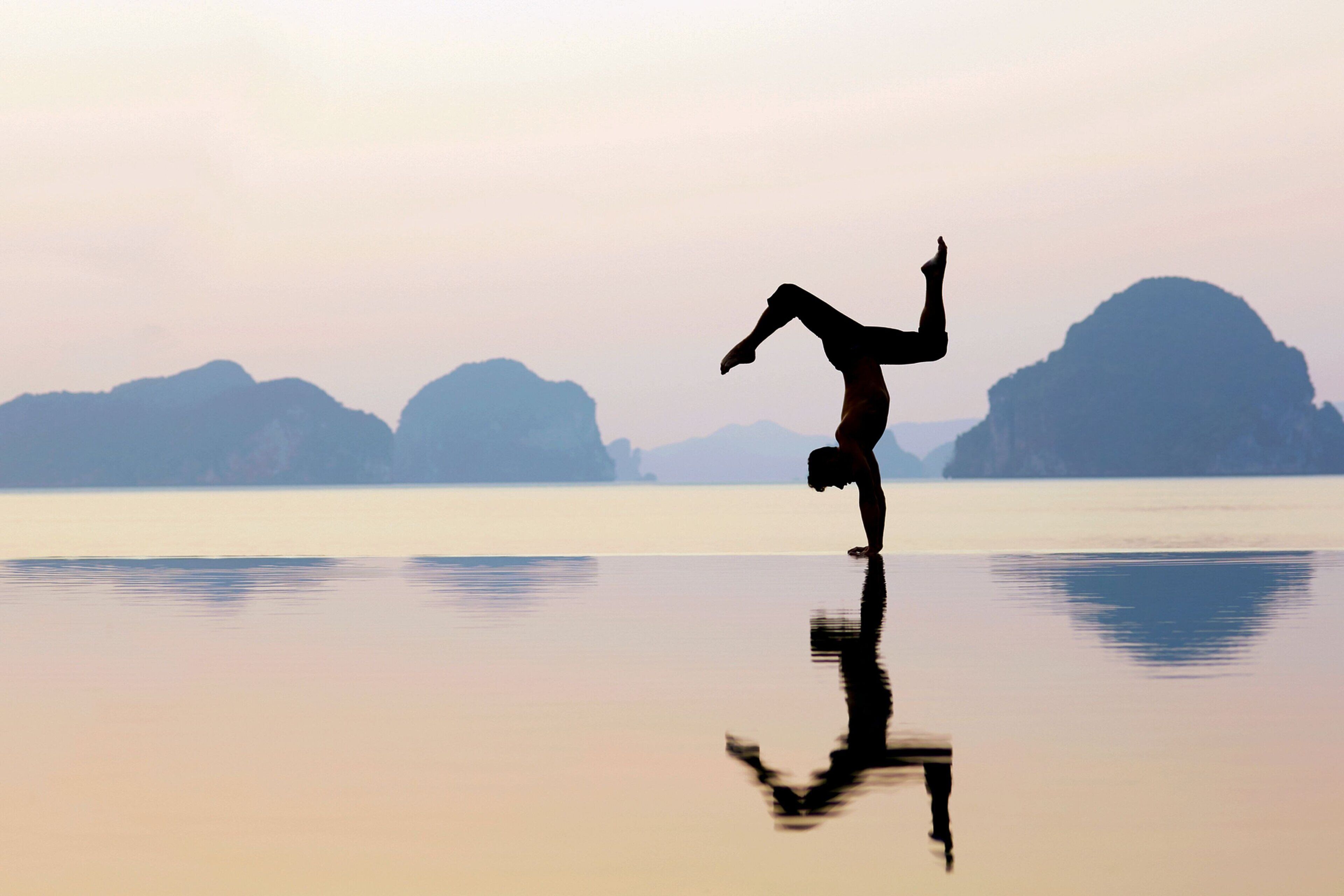 A person performing a handstand on a reflective surface with a calm body of water and distant mountains in the background.