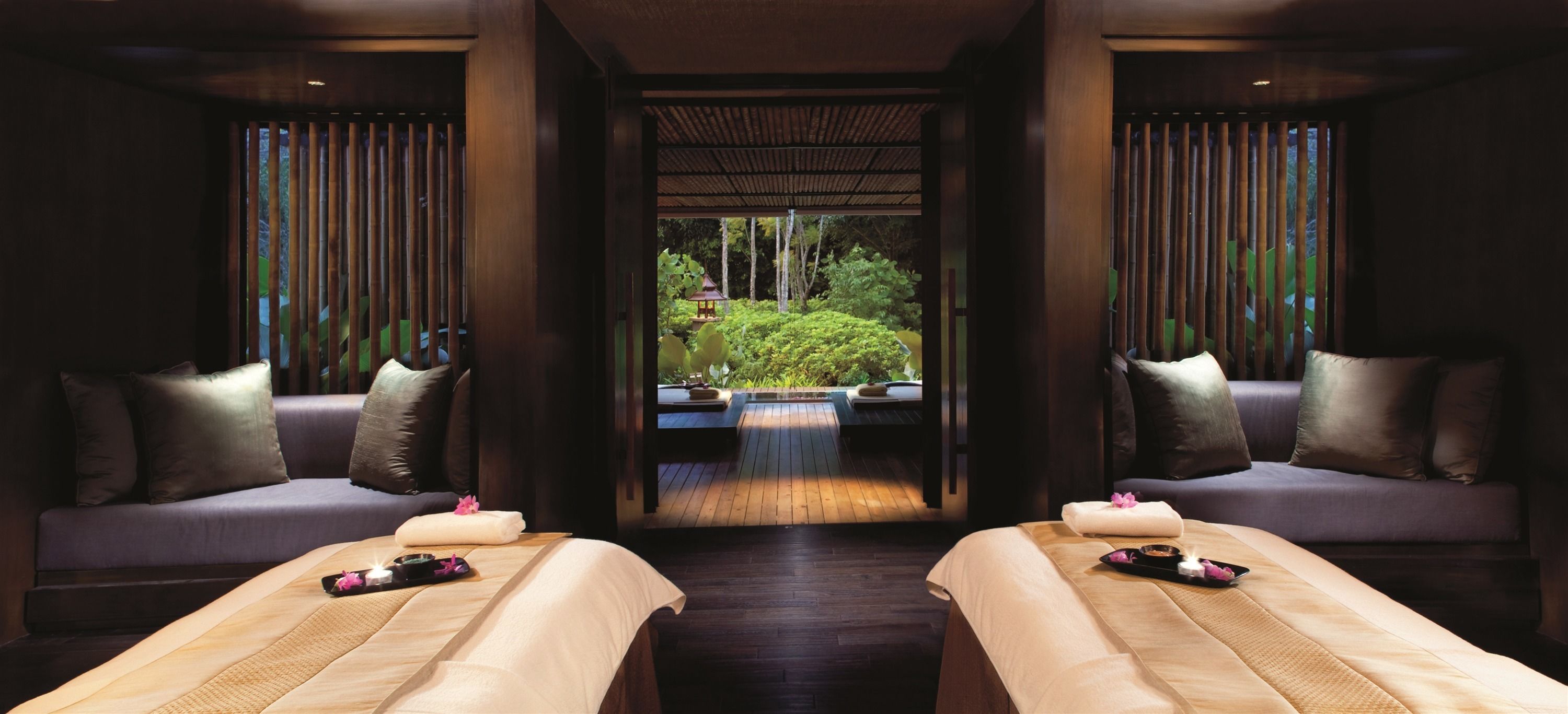 Un cuarto de spa con dos camillas de masaje, suelo de madera y vistas a un jardín a través de una celosía de bambú.