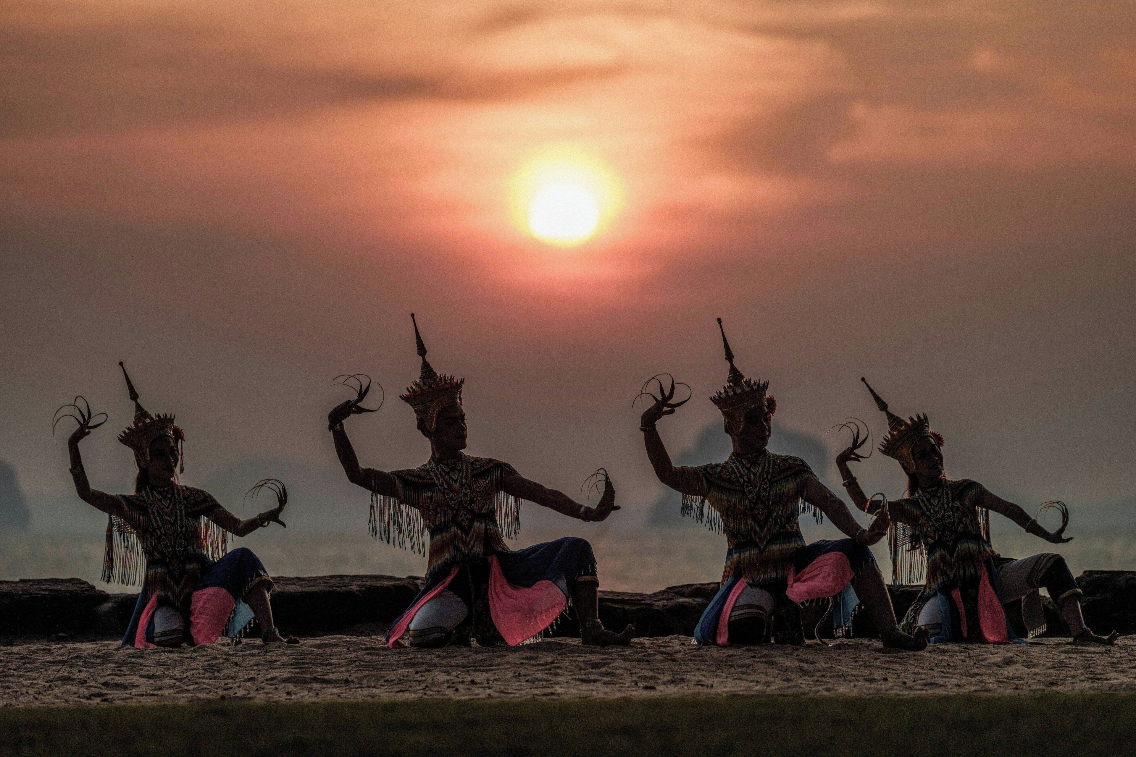 Three individuals in traditional attire seated on a flat surface with a sunset in the background.