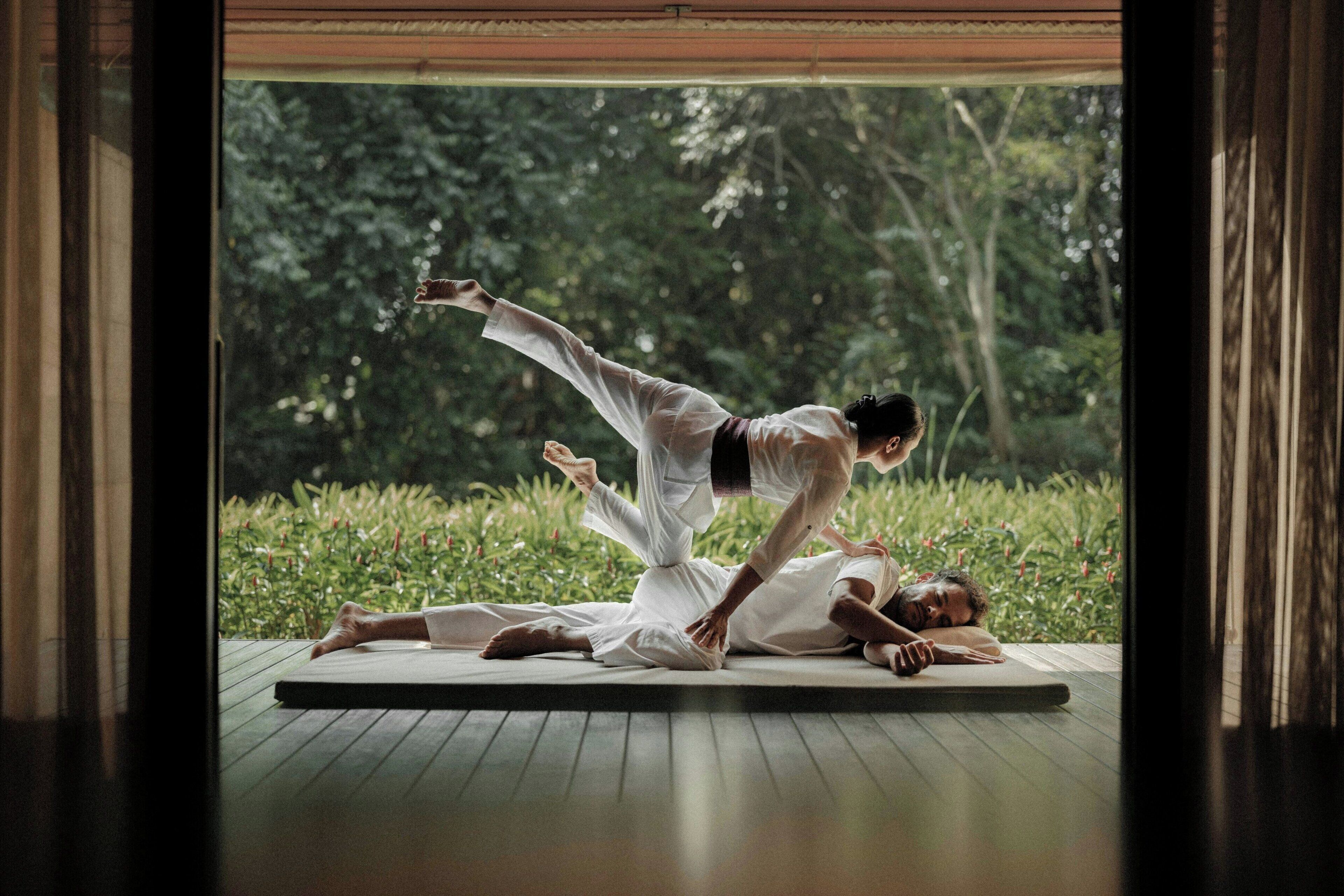 Dos personas practicando yoga sobre una colchoneta en una habitación con vistas a la vegetación.