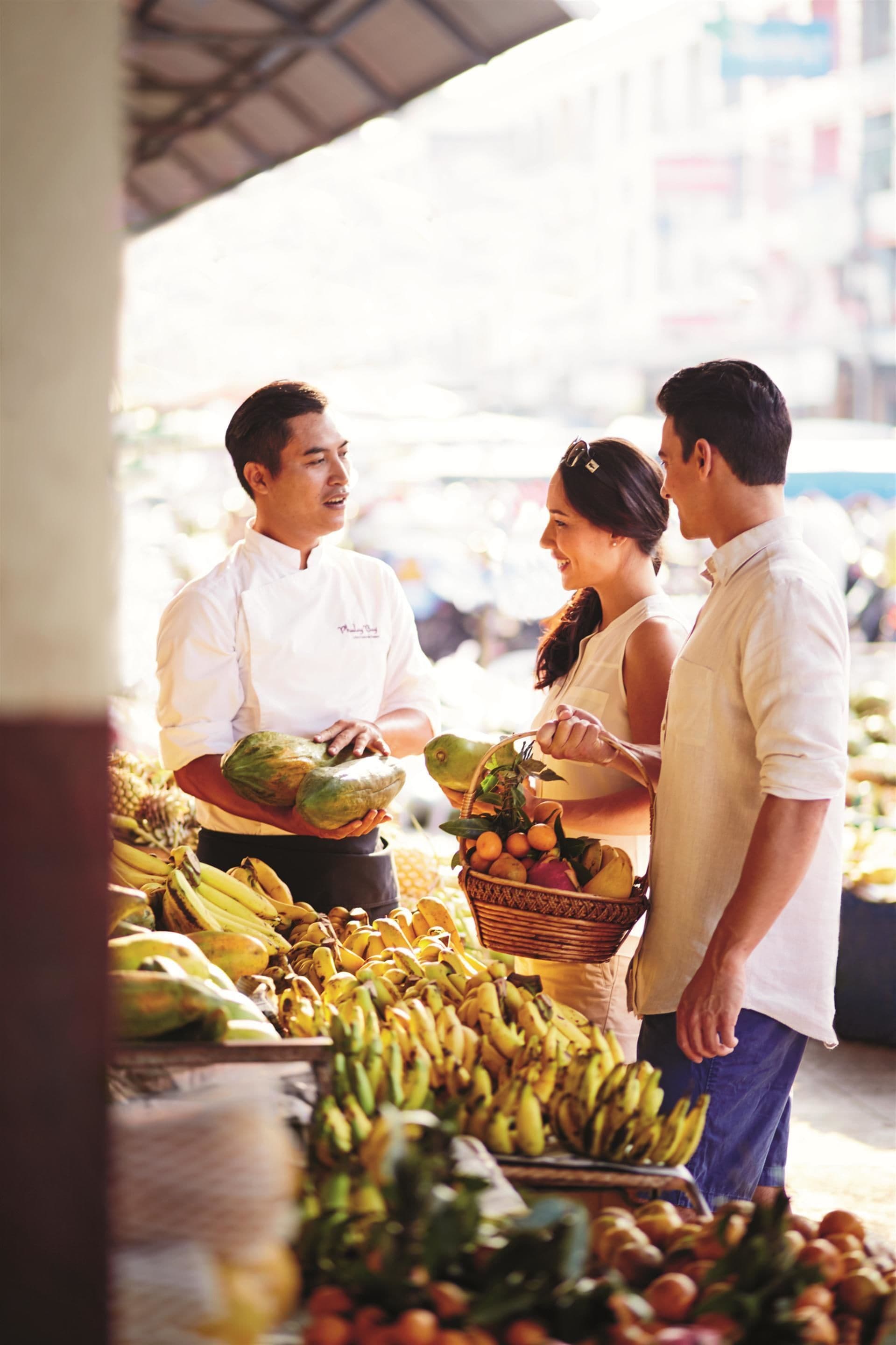 Un étal de marché proposant des produits frais, notamment des bananes et des avocats, et une personne vêtue d'un uniforme de chef.