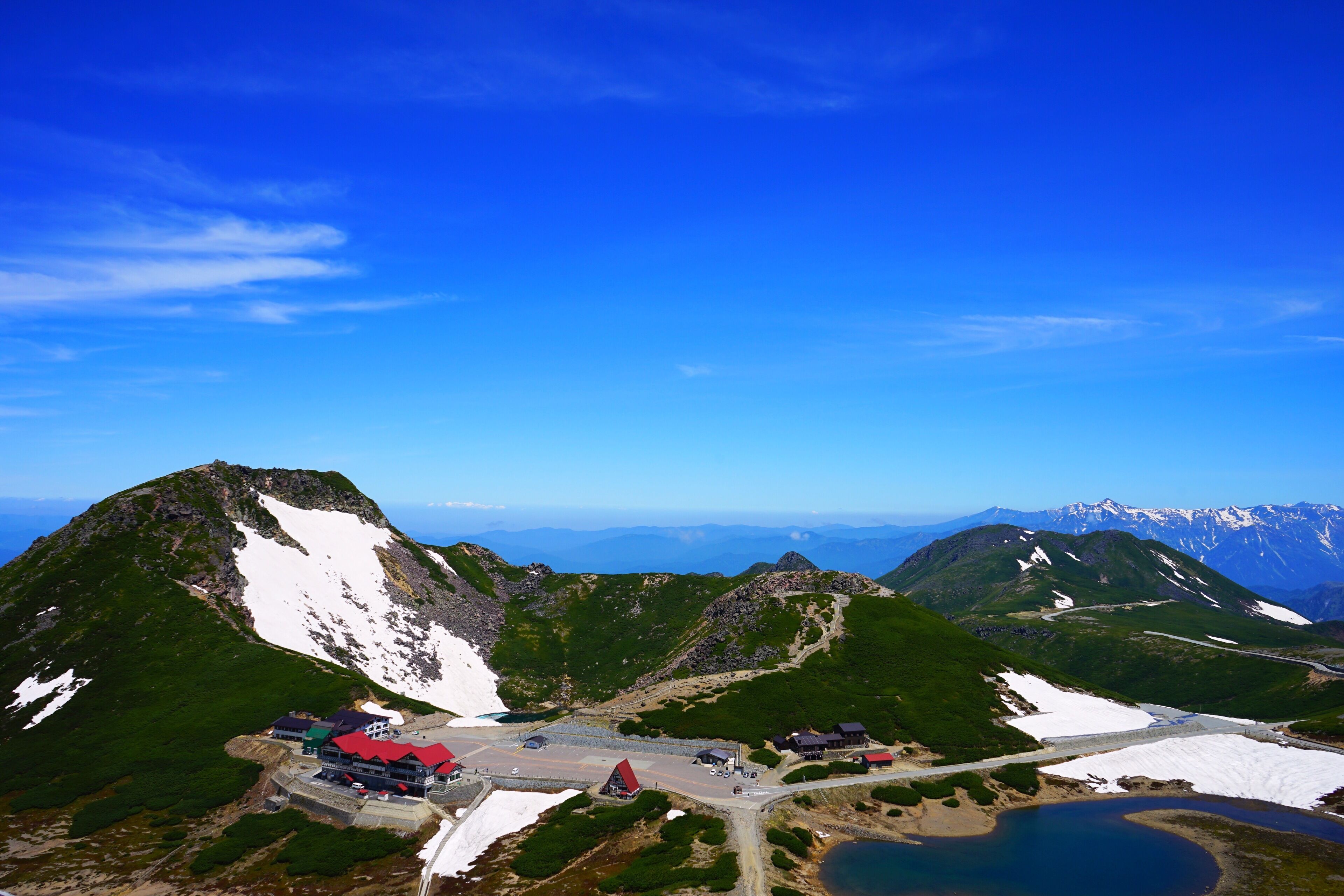 Eine Berghütte mit rotem Dach, umgeben von schneebedeckten Gipfeln und blauem Himmel.