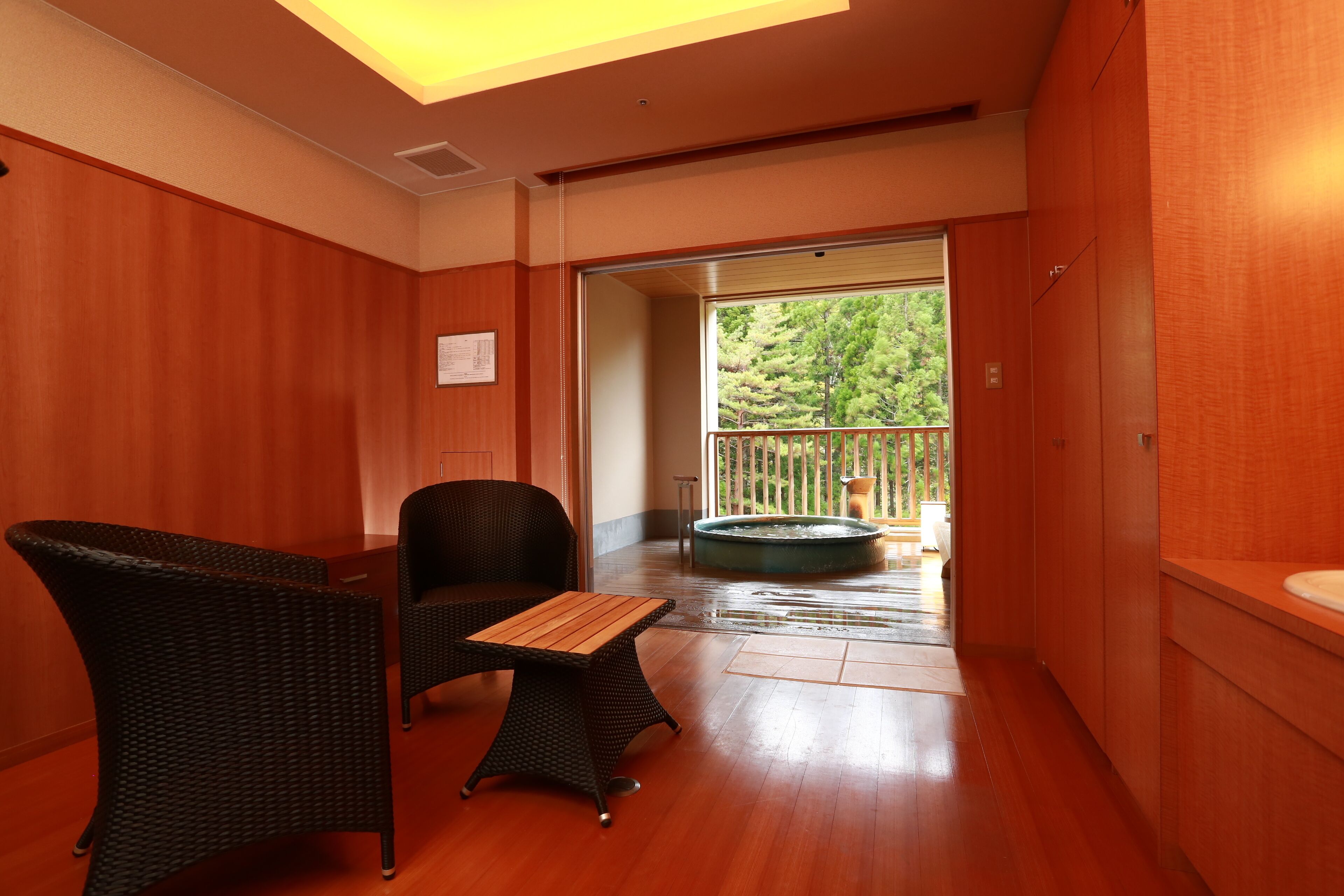 A room with wooden walls, a balcony with a hot tub, wicker furniture, and a view of trees.